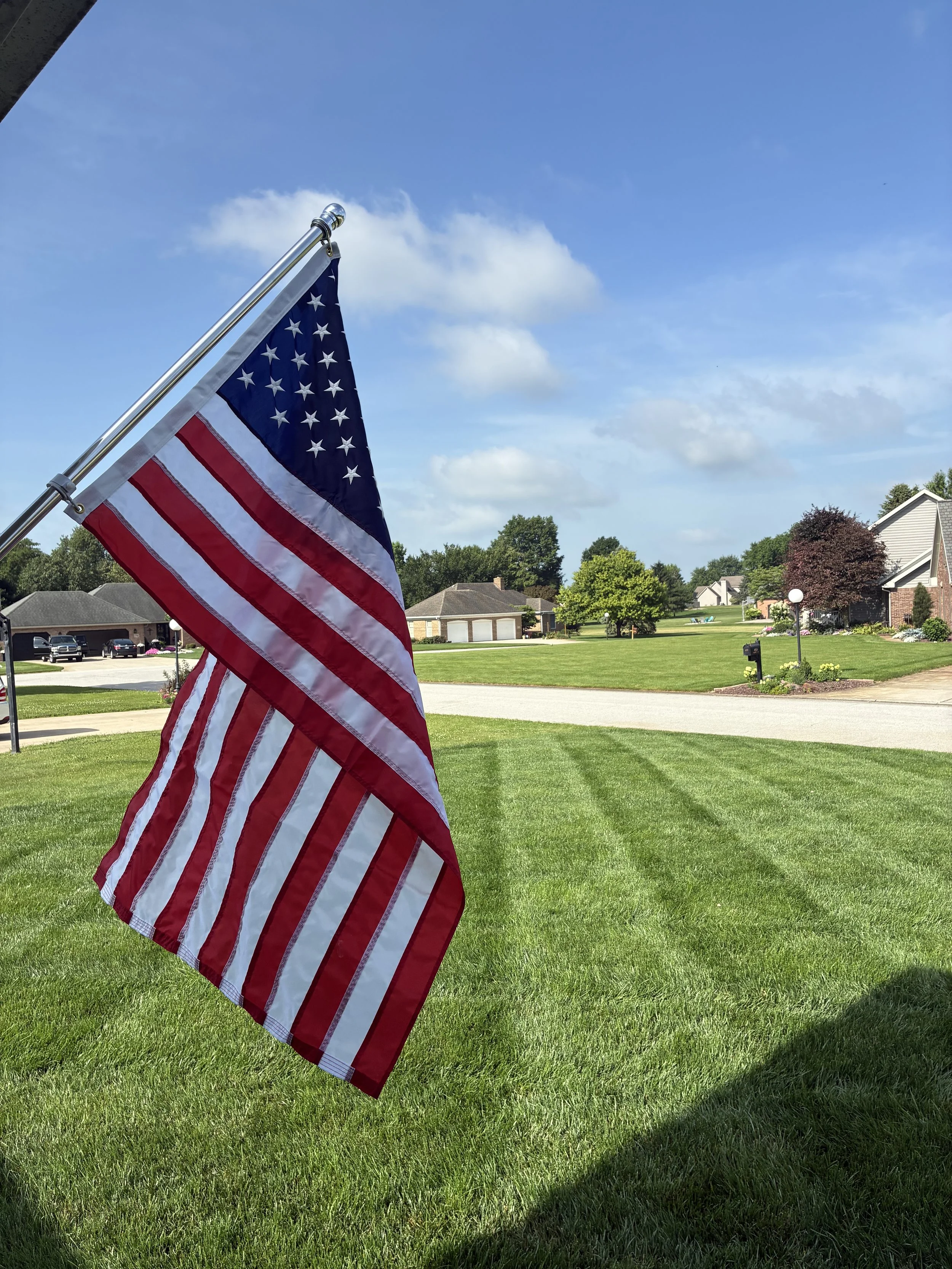 American flag hanging on a flagpole on a residential lawn on a sunny day with a blue sky and some clouds.