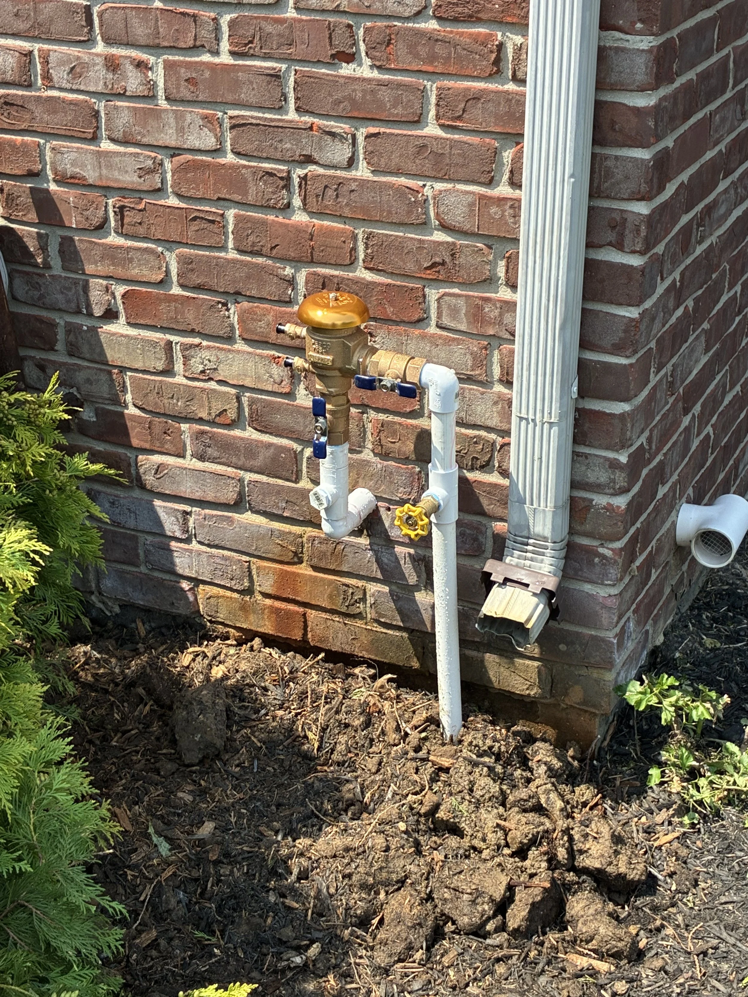 Close-up of a brick exterior wall with a water meter and pipes, with soil and plants in the foreground.