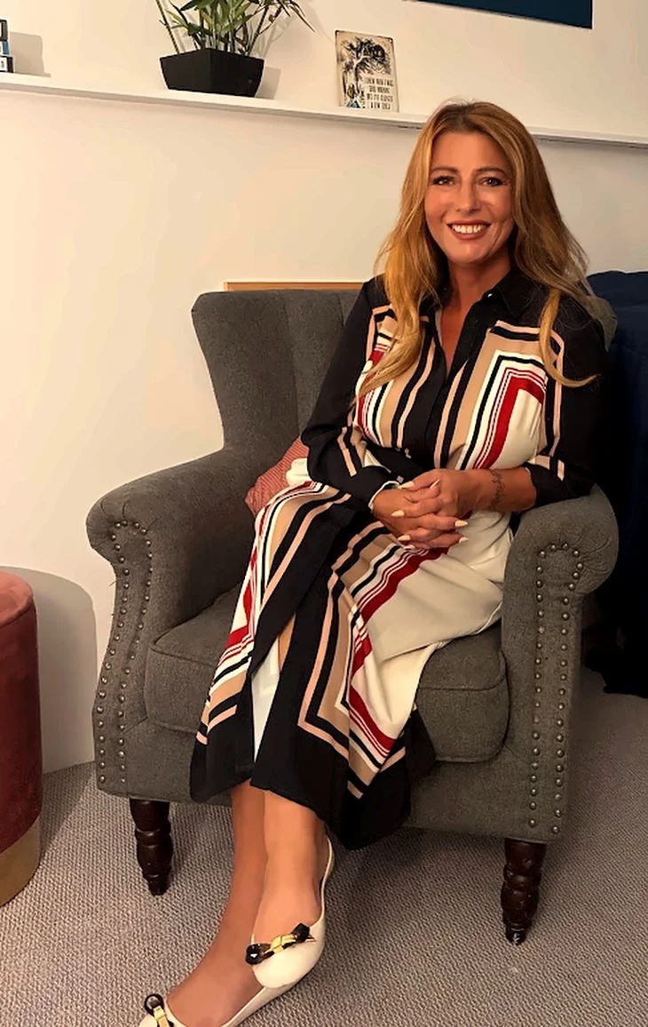 A woman with long, light brown hair wearing a black, white, red, and beige patterned dress, sitting on a gray upholstered armchair with her hands clasped and smiling.