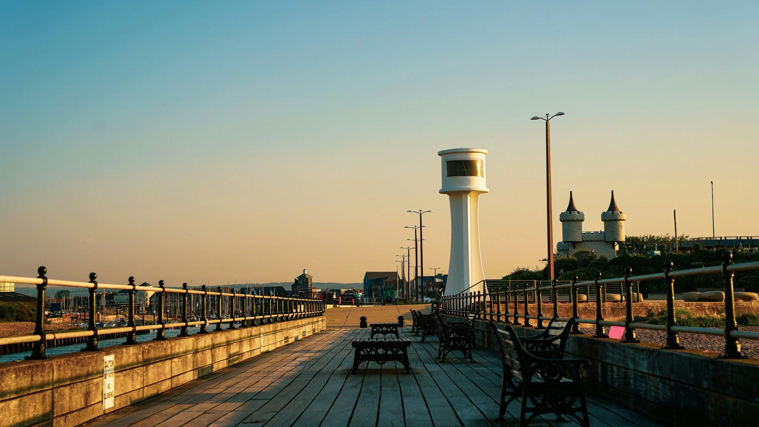 View of a boardwalk with benches, street lamps, and a lighthouse-like tower, near a castle structure, during sunset.