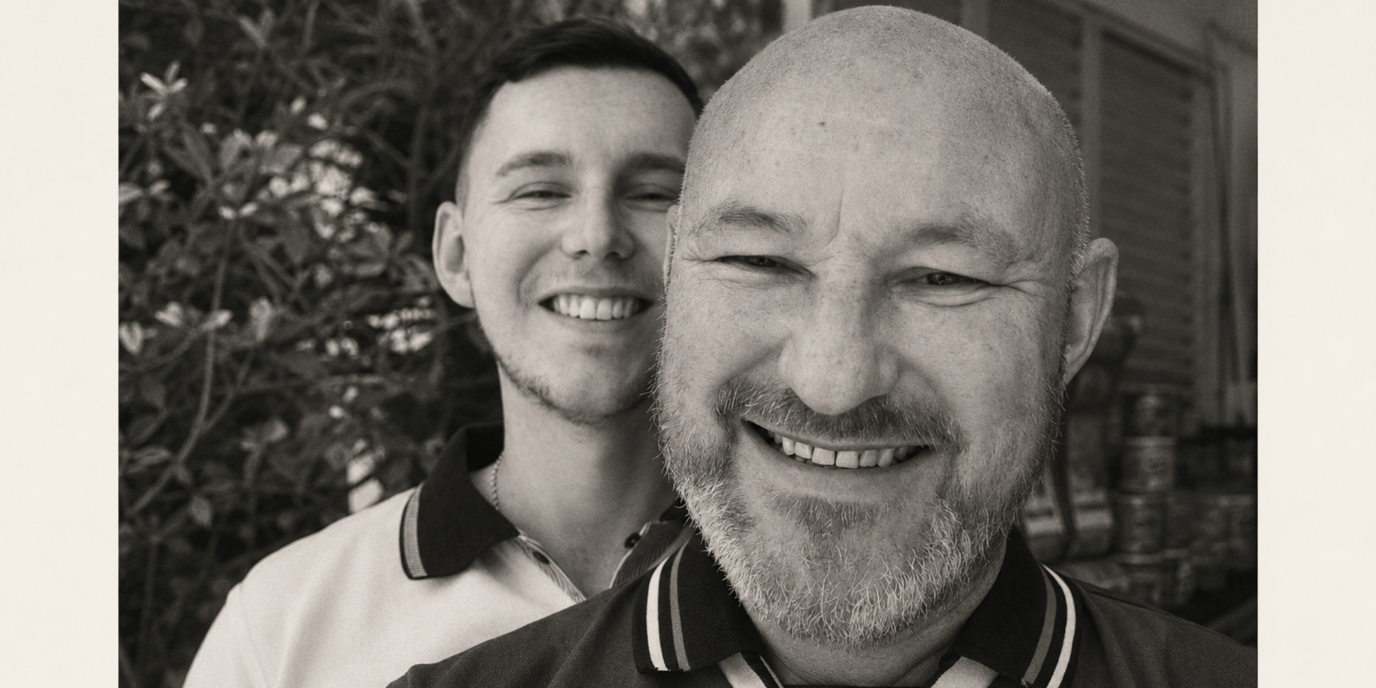 Black and white photo of two smiling men, one with a beard and bald head in front, and another with short hair and a mustache in back, outdoors with blurred foliage in background.