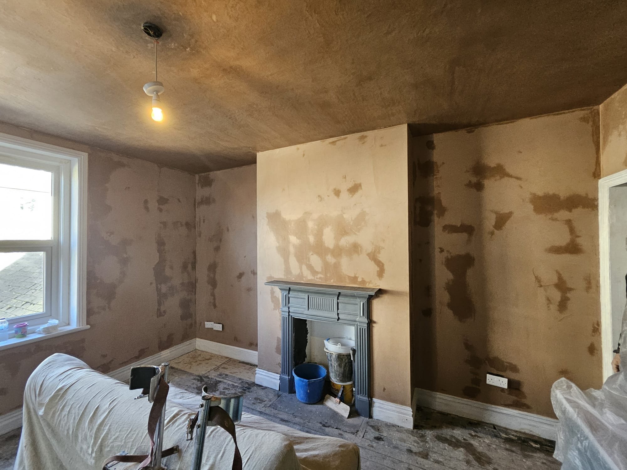 A room under renovation with unfinished walls, a window, and household items covered with a sheet, including a chair and buckets near a fireplace.