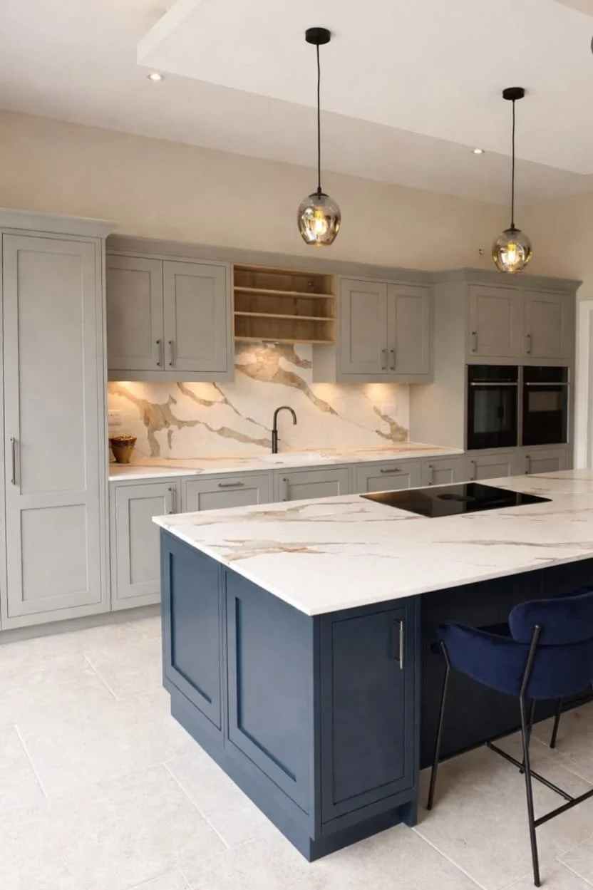 Modern kitchen with gray cabinetry, marble backsplash, and island with a blue base. Pendant lighting above the island, built-in double ovens, and a black cooktop on the island. There is a chair with navy velvet upholstery beside the island.
