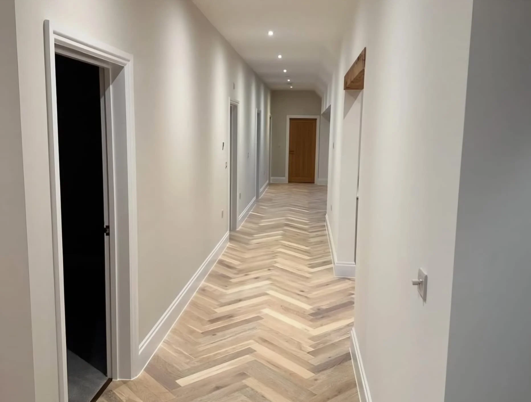 Modern indoor hallway with white walls, wooden floor, and doors along the sides. Recessed ceiling lights illuminate the space. One door appears to be open to an elevator.