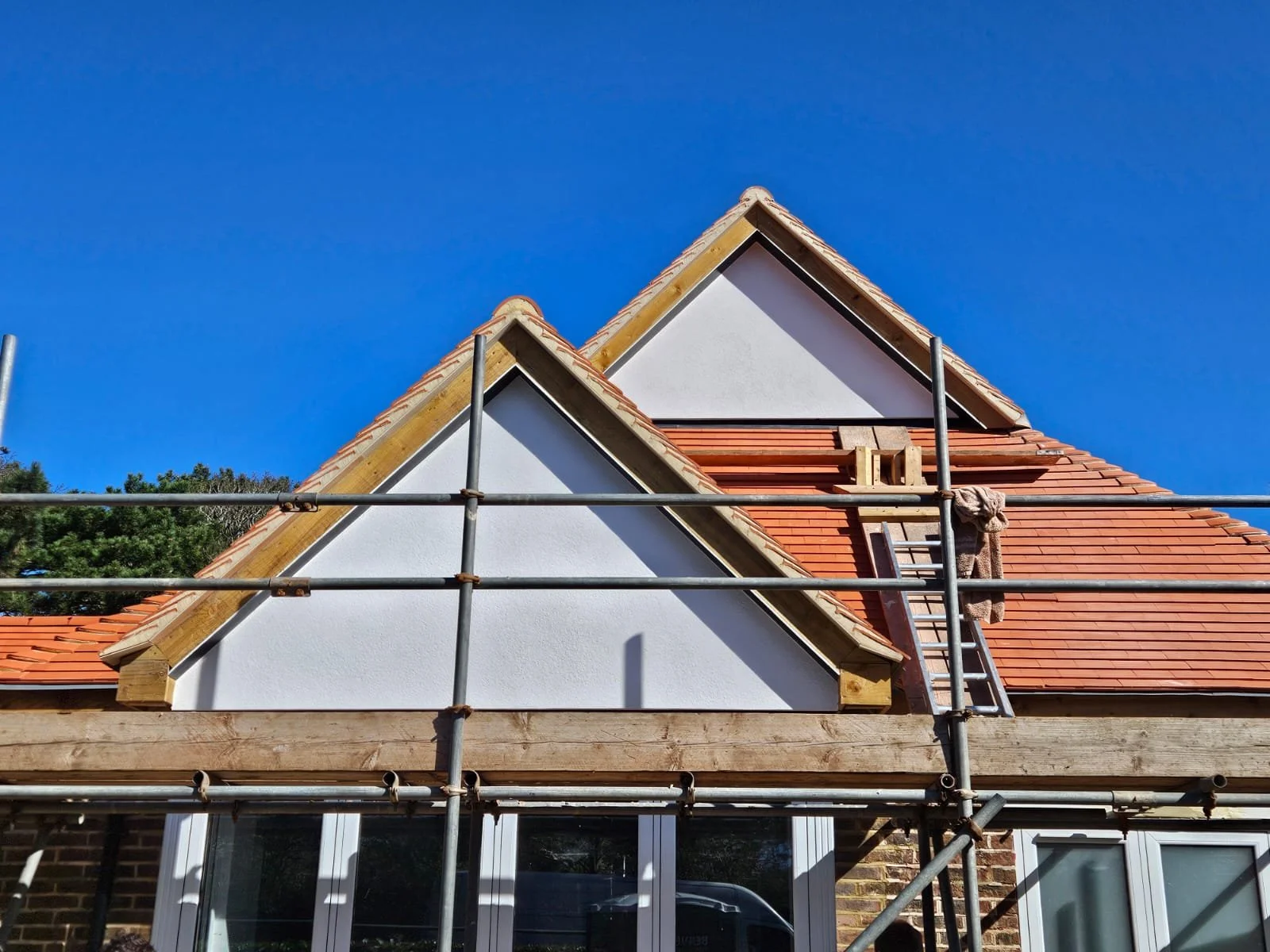 House under construction with scaffolding, showing a steep roof with red tiles, white walls, and windows at the bottom. There is a ladder leaning against the roof, and a towel hanging on the ladder. Blue sky in the background.