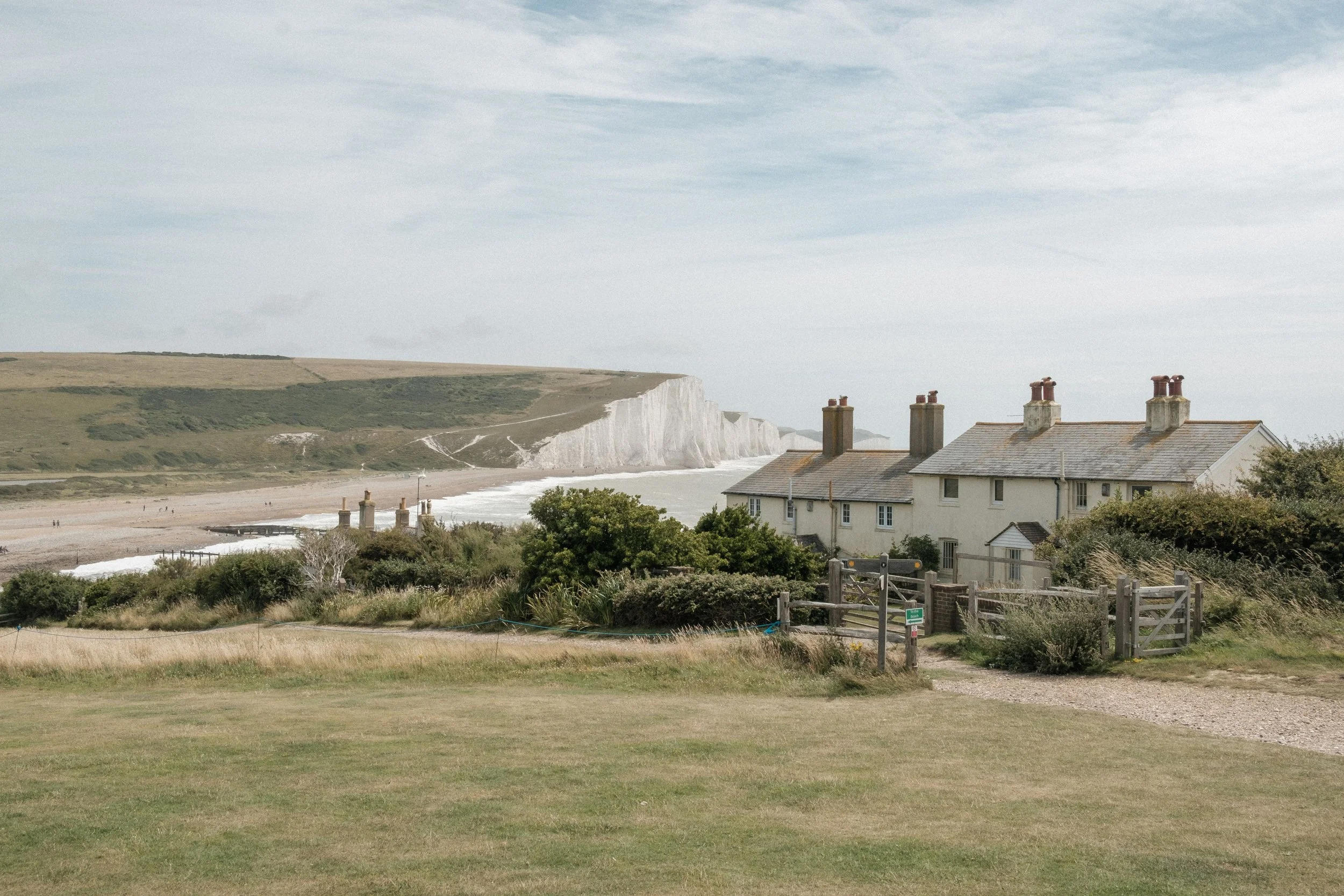 Scenic view of white chalk cliffs along a beach, with residential houses in the foreground and a grassy area, under a cloudy sky.
