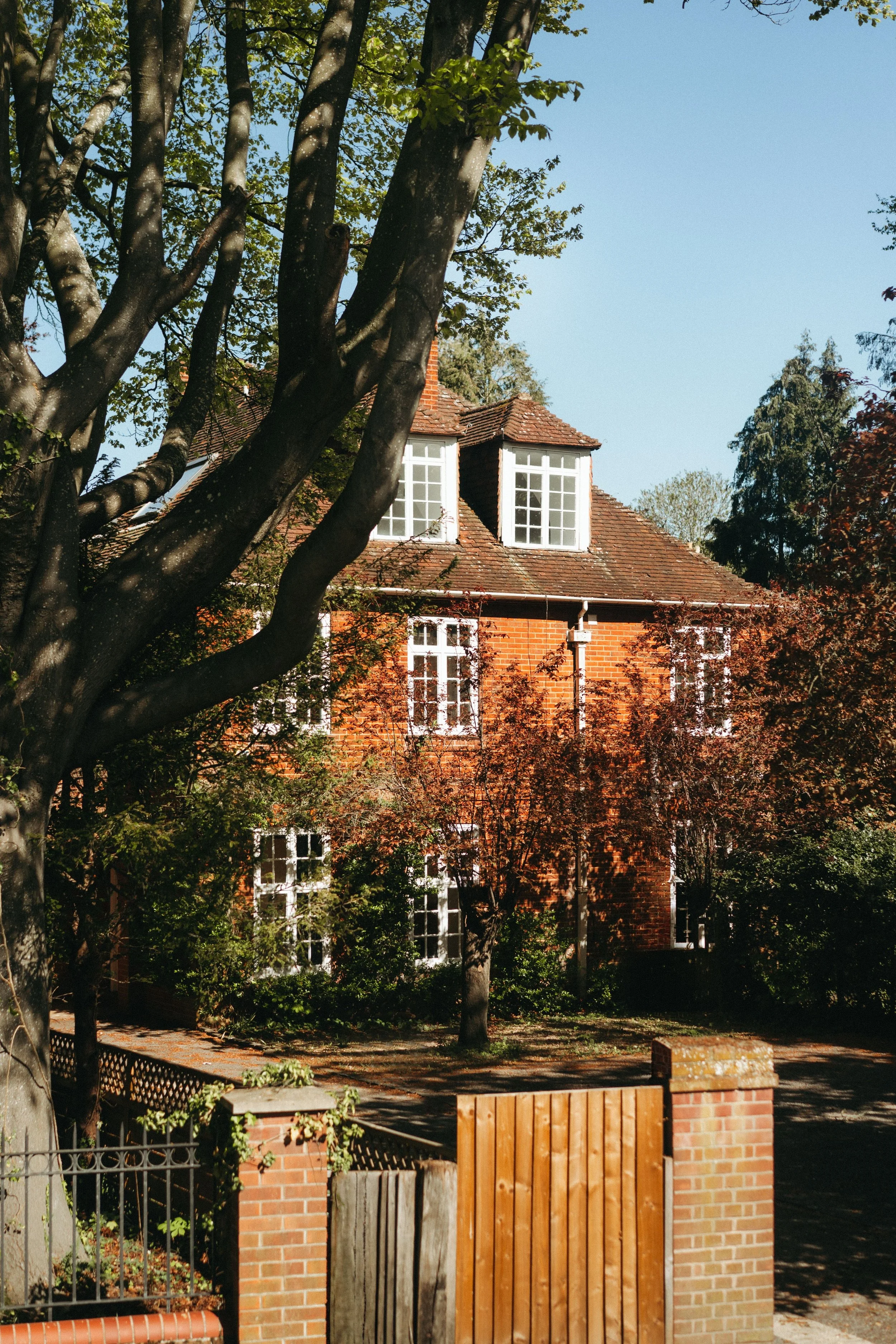 A brick house with a tiled roof, two dormer windows, surrounded by trees and a small yard, with a wooden gate and brick fence in the foreground.
