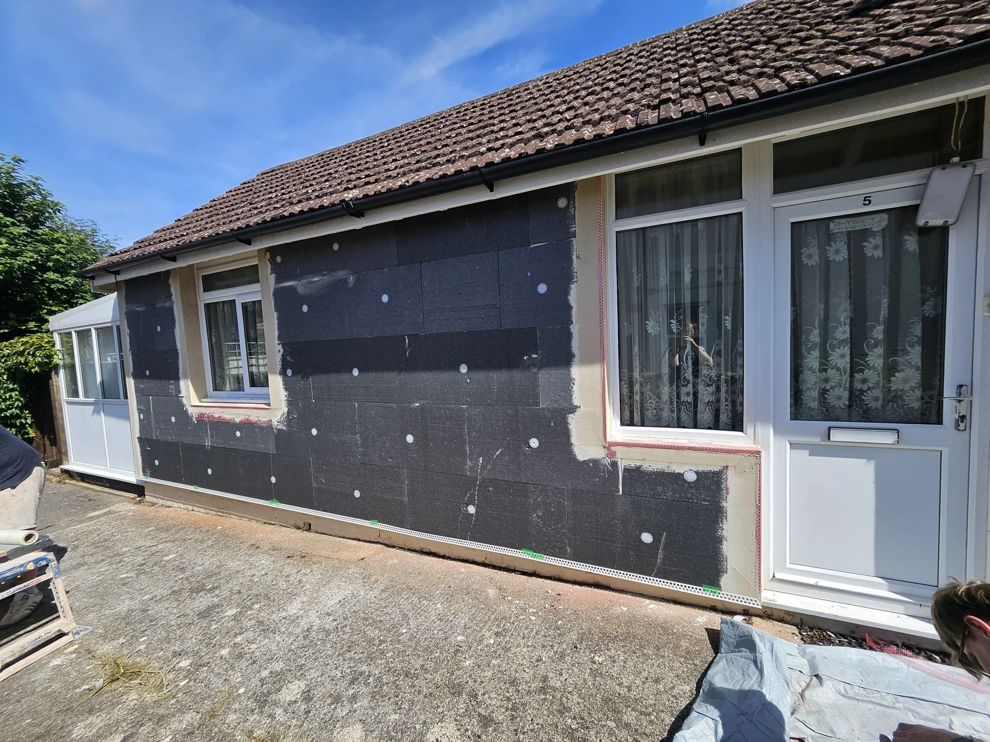 House exterior undergoing renovation with black insulation board on wall, white window and door frames, and a tiled roof. Construction tools and materials are visible on the ground outside.