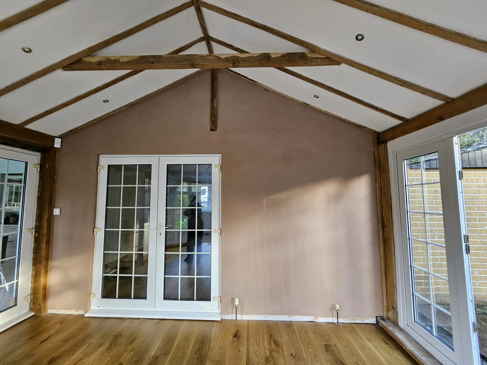 Interior of a room with a vaulted ceiling, showing wooden beams and joists, two closed French doors with glass panes, a brown wall, and a hardwood floor.