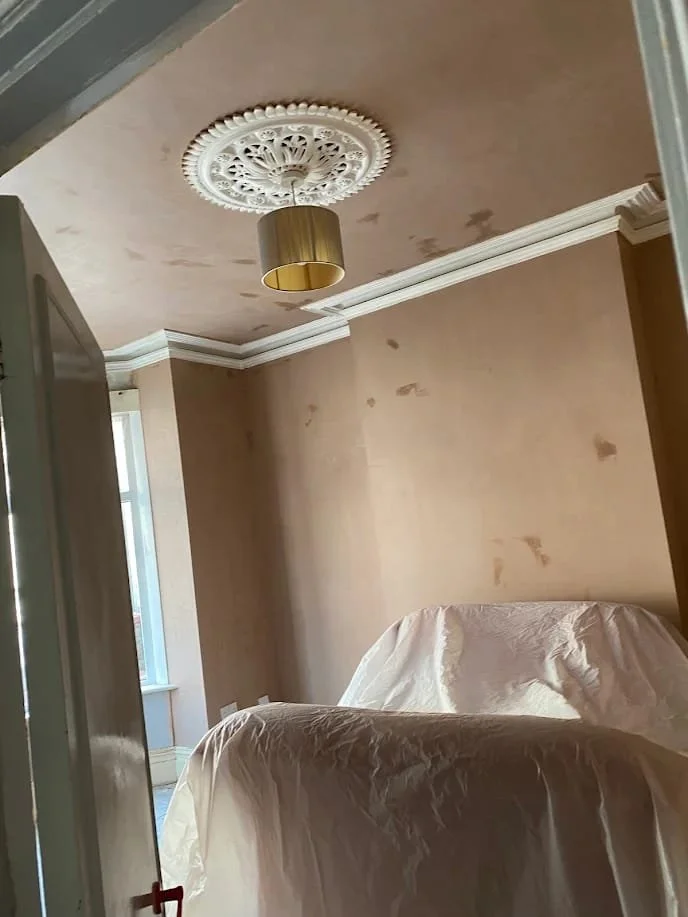 Interior of a room under renovation with beige walls, white crown molding, a ceiling medallion, and a hanging light fixture. Furniture covered with protective cloth is visible in the foreground.