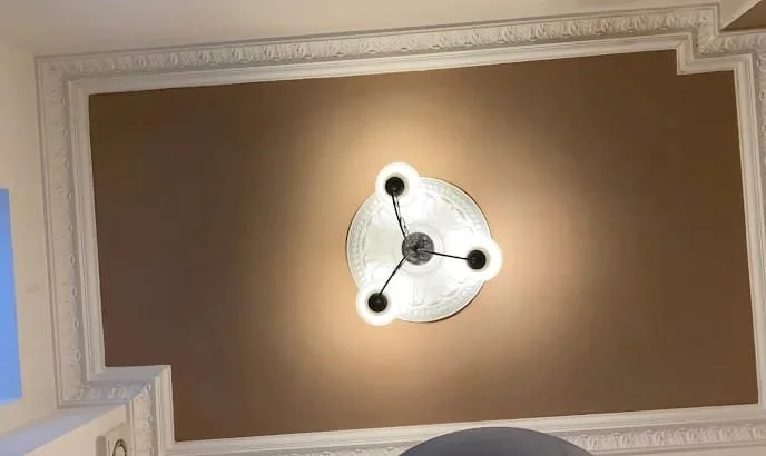 Ceiling light fixture with three circular lights connected by black rods, mounted on a decorative white ceiling medallion in a room with beige walls.