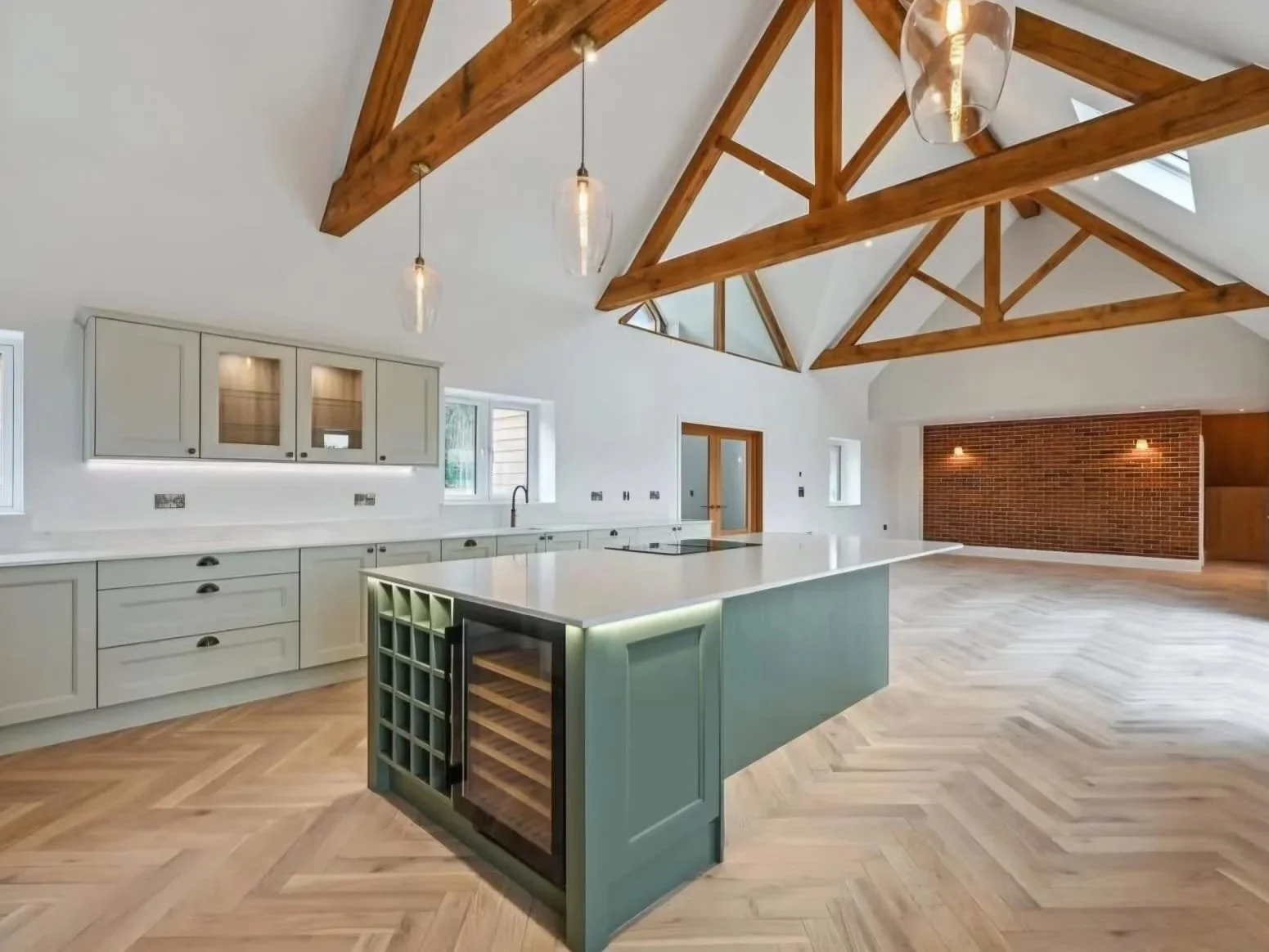 Modern kitchen with a large island, light gray cabinets, wooden vaulted ceiling with exposed beams, hardwood herringbone floor, brick wall, and pendant lights.