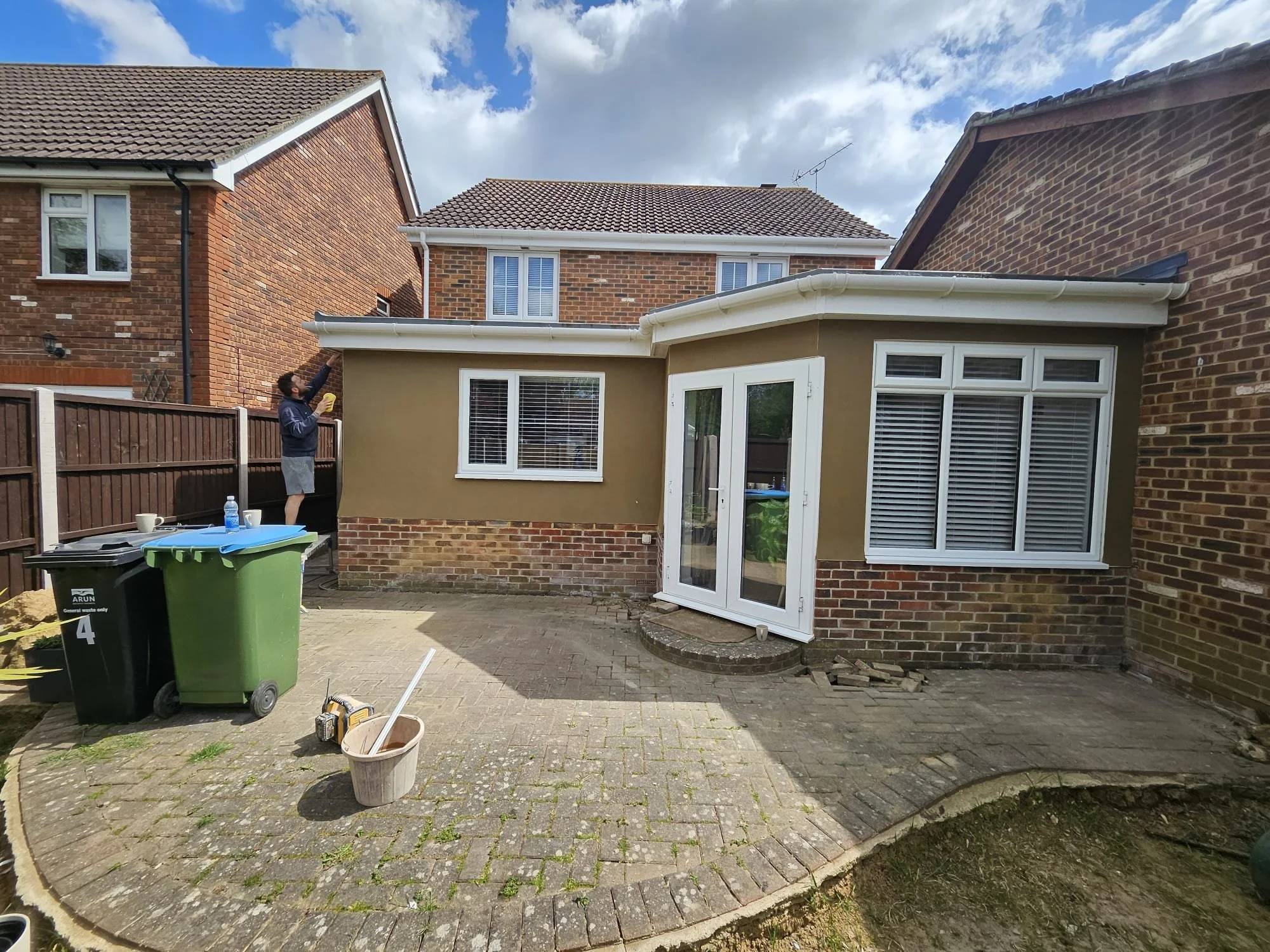A man is cleaning the roof eaves of a house in a backyard. There are green and black trash bins, gardening tools, and a bucket on the ground, with a brick paved patio area surrounded by a wooden fence. The house has brick walls, white window frames, and a conservatory attached to the back.
