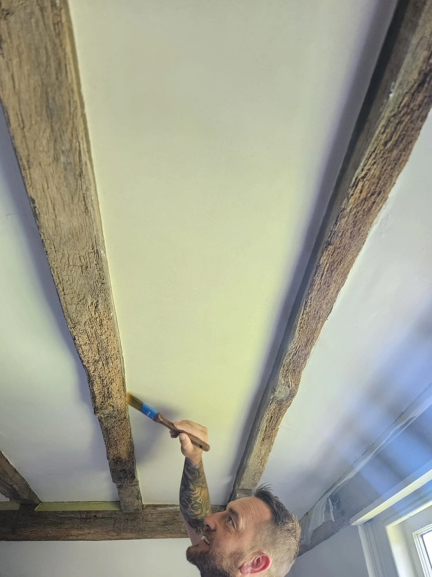 A man with tattoos on his arm is painting a white wall with a paintbrush. The ceiling is decorated with wooden beams.