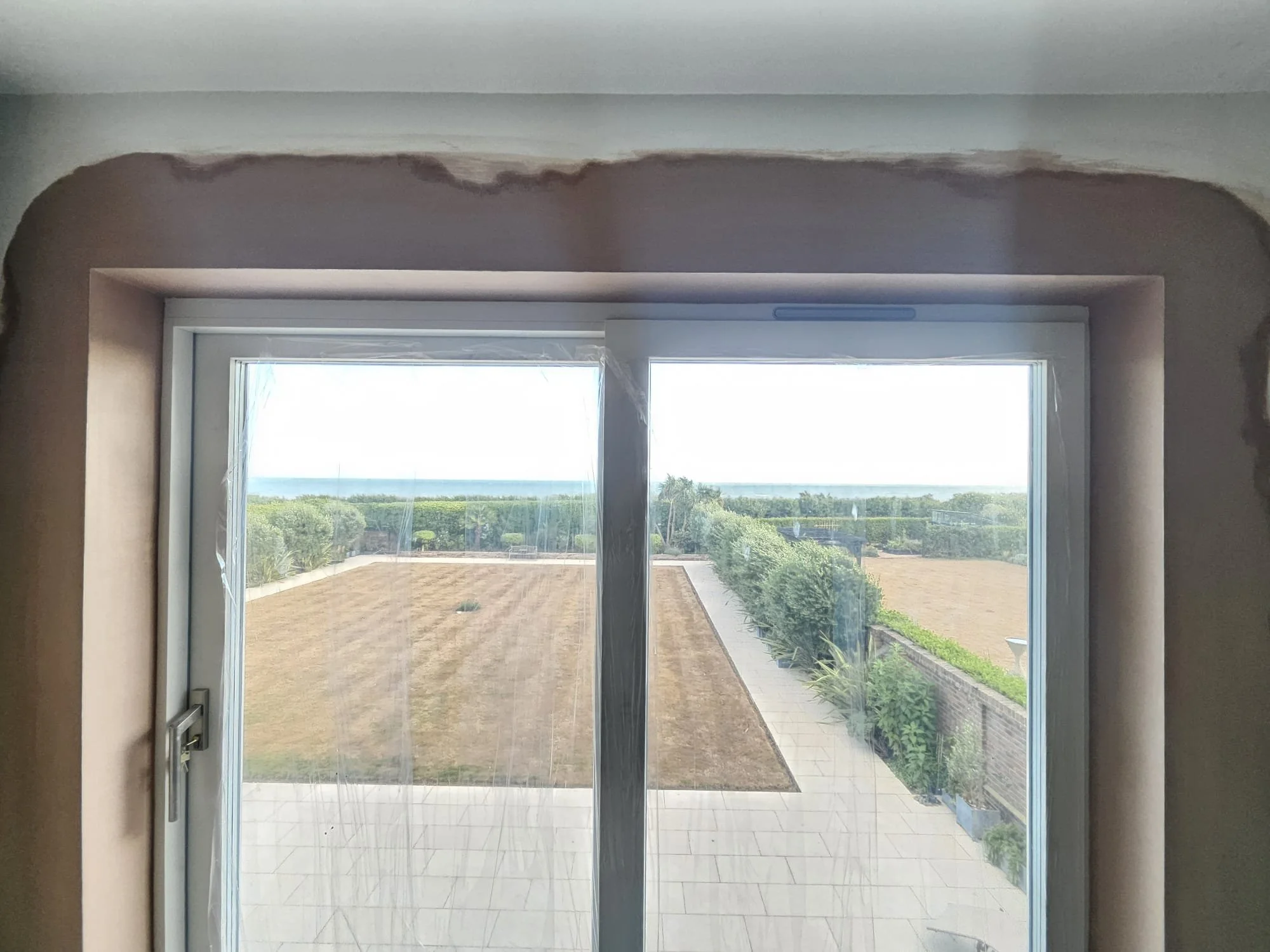 View through a sliding glass door showing a backyard with a dirt or sand area, a walkway, lush greenery, bushes, and trees, with a clear sky and distant ocean horizon in the background.