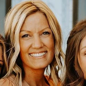 Smiling woman with long, blonde highlights in hair, surrounded by two partially visible people, indoors.