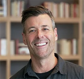 Smiling man with dark hair and gray beard in front of bookshelves.