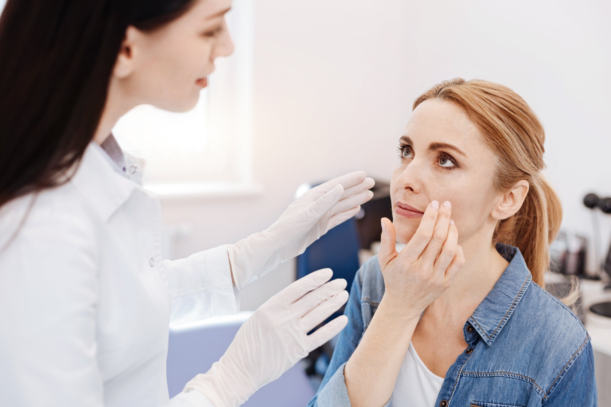 A healthcare professional wearing gloves examines a woman's face during a medical consultation or skincare assessment in a clinical setting.