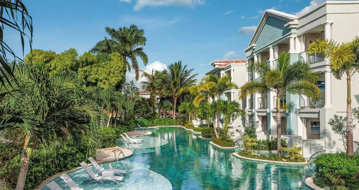 A wooden poolside house with a steep roof, large windows, and a deck area with lounge chairs and cushions. There is a swimming pool in the foreground with a child playing in the water and an adult nearby. A palm tree is leaning against the house. The scene appears to be at sunset with warm lighting.