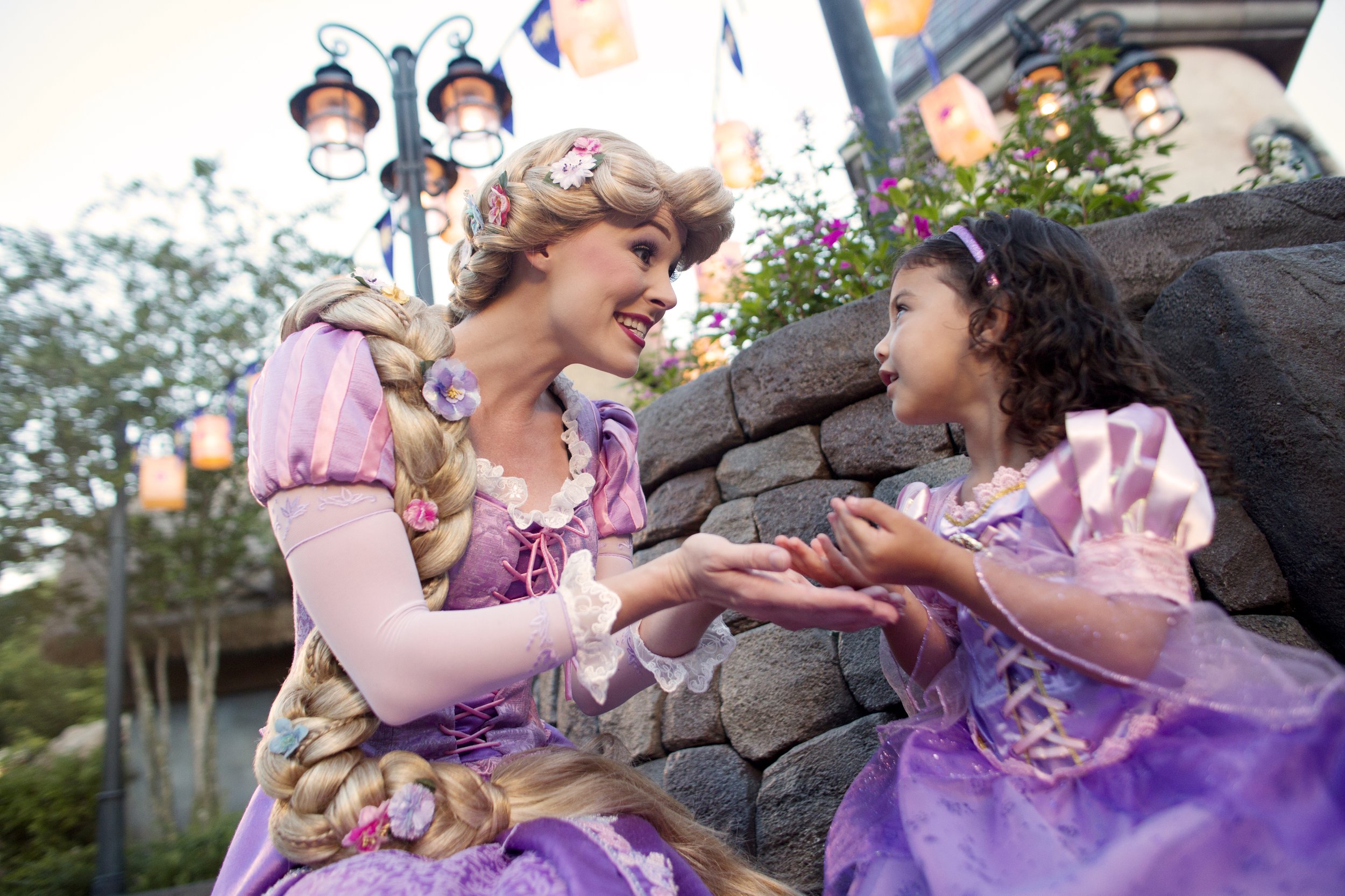 A woman dressed as Rapunzel from Disney's Tangled, with long braided hair decorated with flowers, smiling at a young girl in a purple princess dress. They are outside by a stone wall, with hanging lanterns and flags in the background.