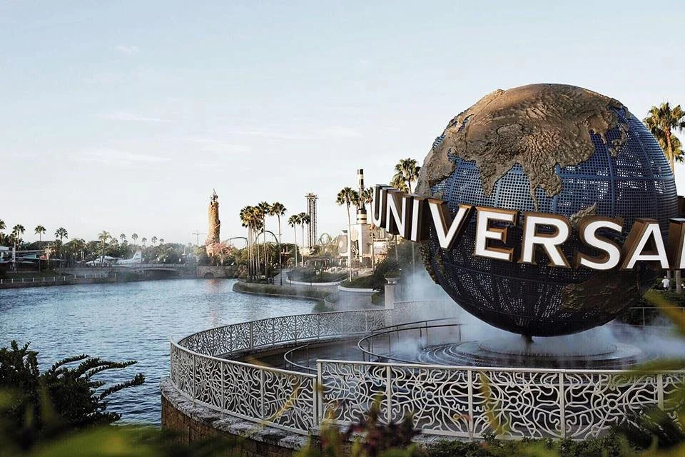 Universal Studios globe in front of a water lagoon with palm trees, iconic rides, and a partly cloudy sky in the background.
