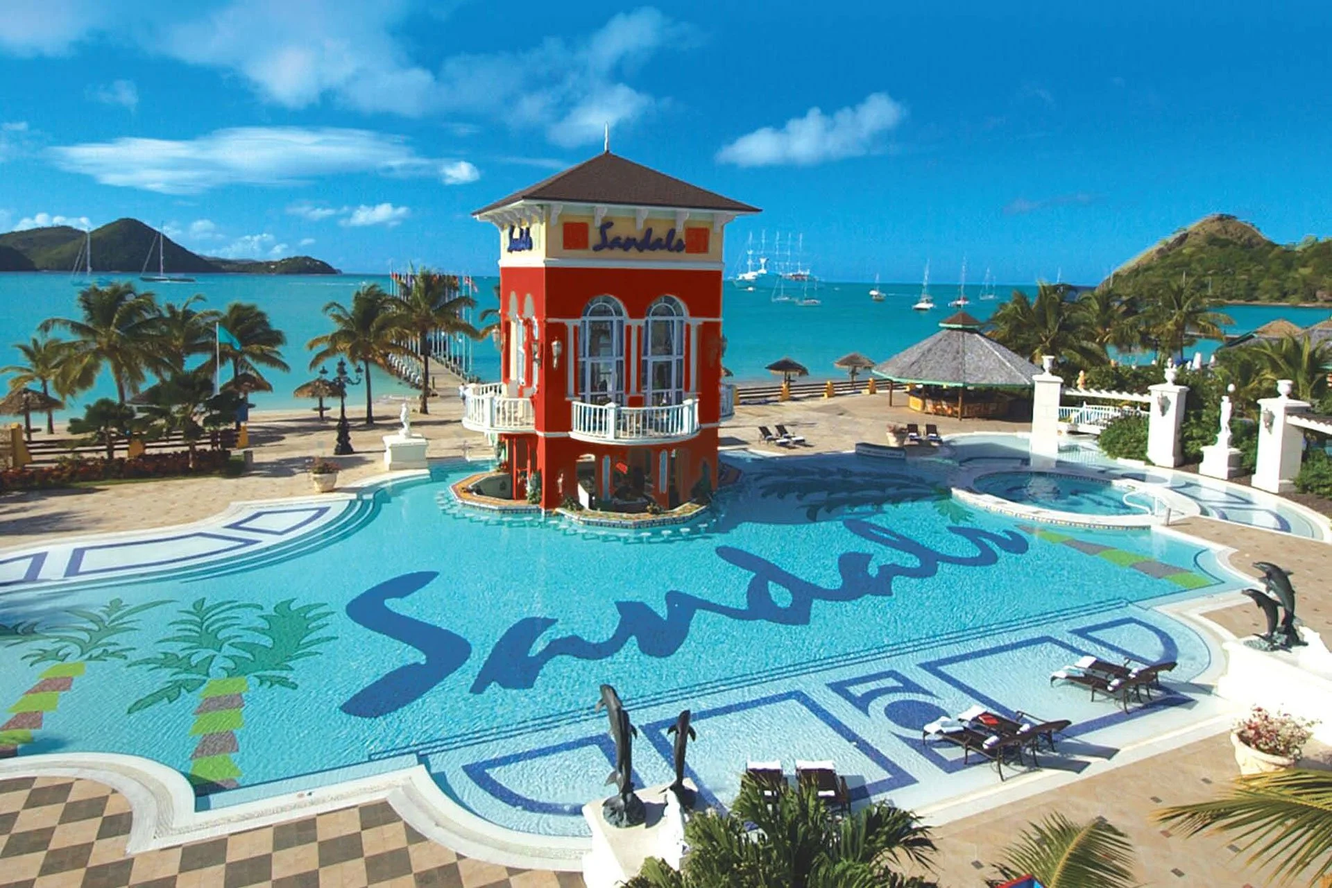 A resort pool area with a red tower labeled 'Sandals' by the sea. The pool has dragon designs, surrounded by lounge chairs and palm trees, with sailboats on the ocean and green hills in the background.
