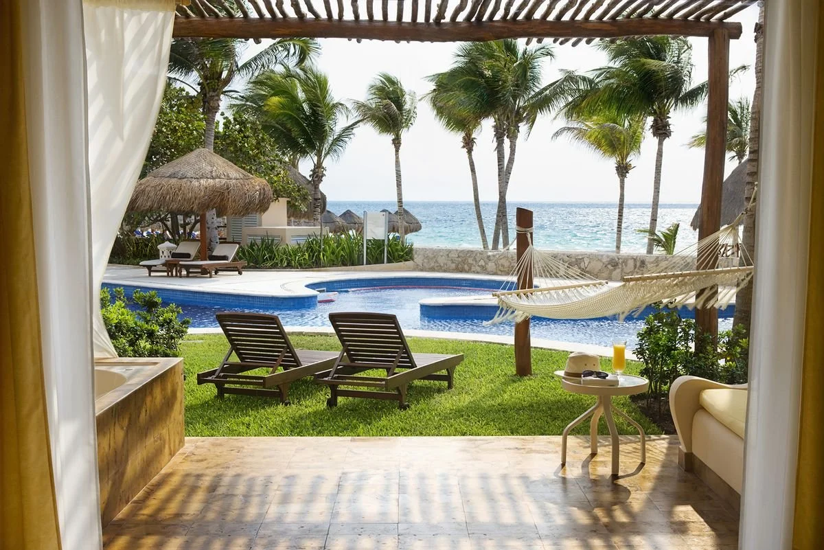 View of a swimming pool area with lounge chairs, a hammock, and palm trees overlooking the ocean on a sunny day