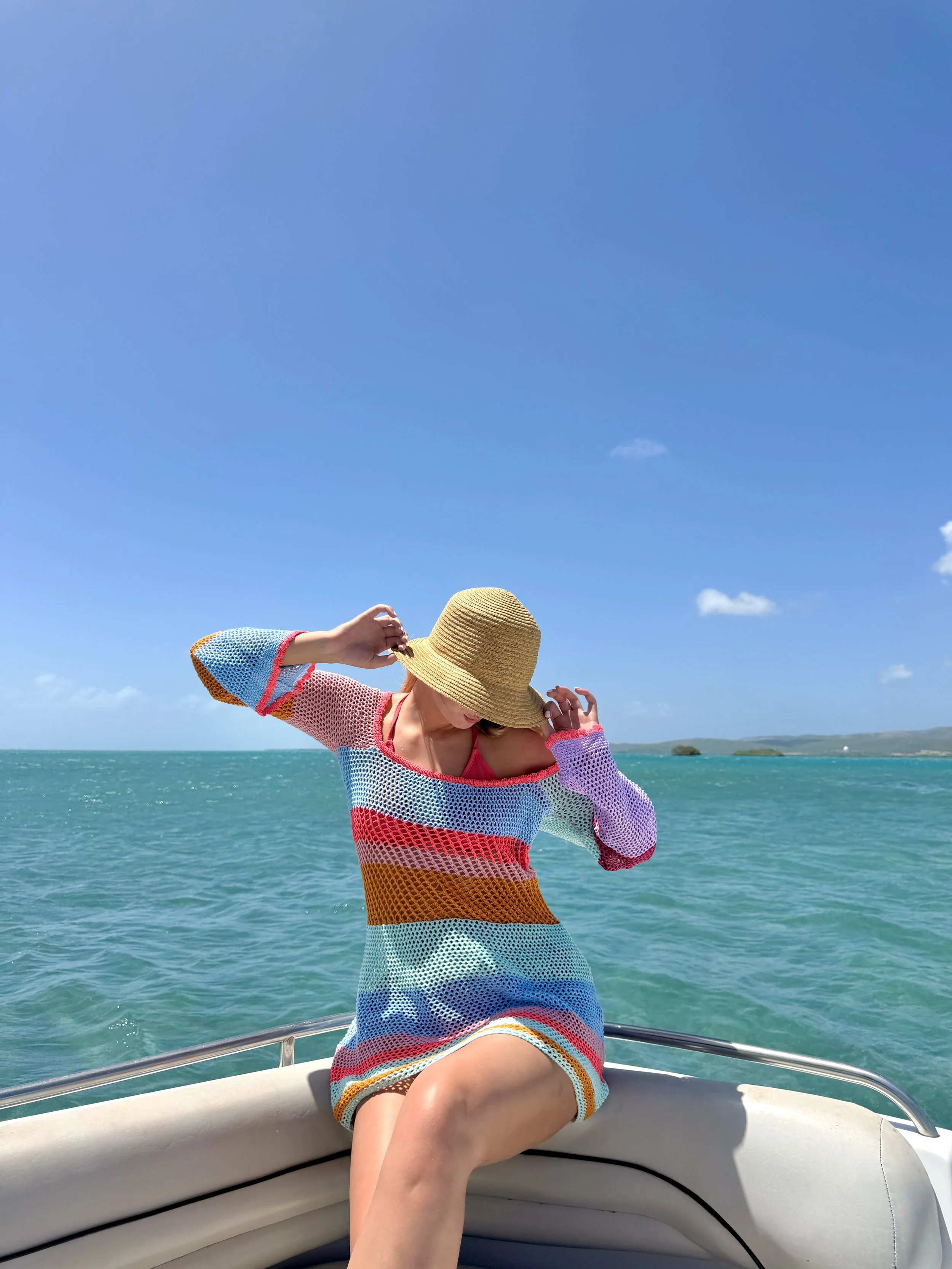 A woman in a colorful striped crochet dress and a wide-brimmed straw hat sitting on a boat, adjusting her hat while near the ocean under a bright blue sky with a few clouds.