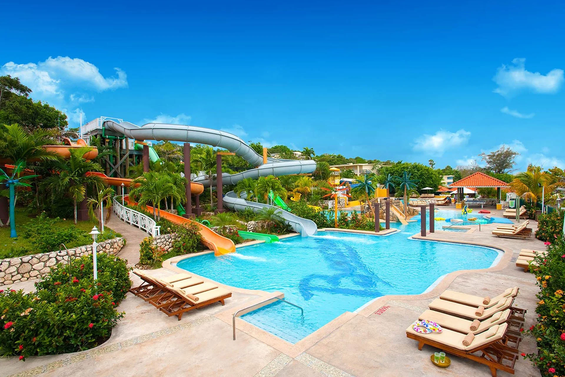 Large outdoor water park with multiple water slides, lazy river, and pool area surrounded by lounge chairs and lush tropical plants under a bright blue sky.
