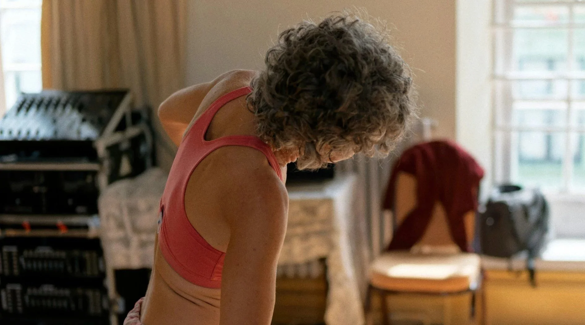 An elderly woman with curly gray hair wearing a pink sports bra, looking downward in a room with natural light coming through window blinds. Background includes chairs, a bookshelf, and a red bag.