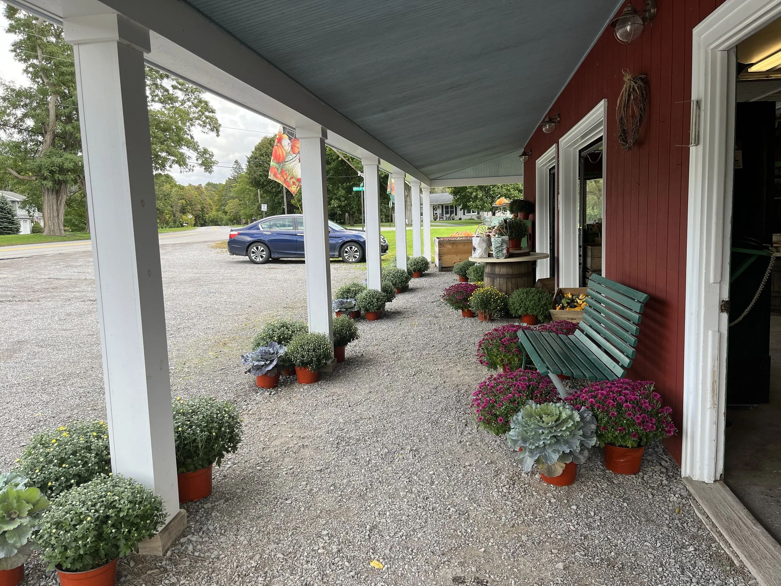 A porch with a red wall and white trim, decorated with potted flowers and plants. A green slatted bench is positioned on the right side, and a table with additional plants and flower arrangements is visible. The porch is supported by white columns, a