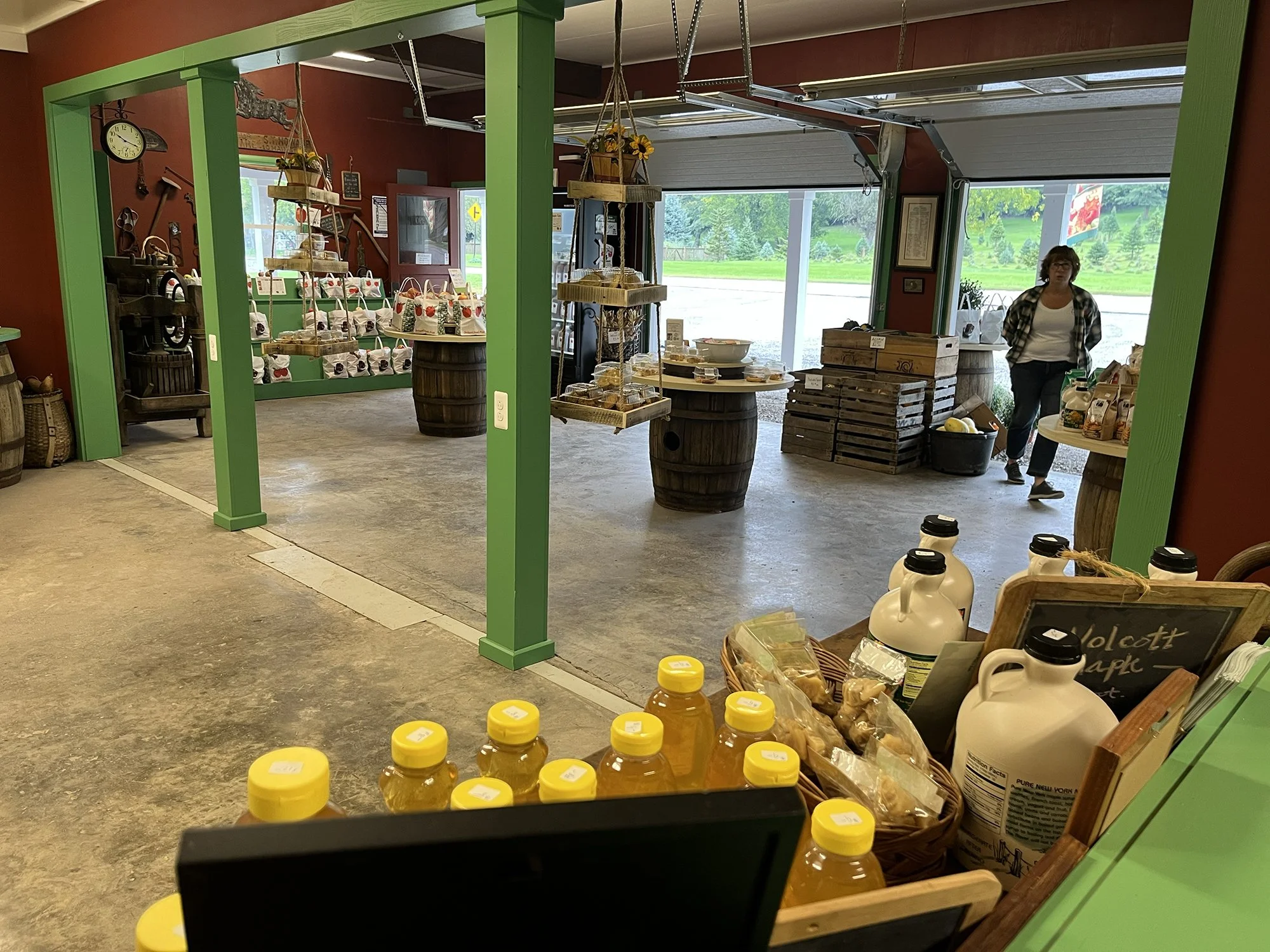 Interior of a farm store with green framing, displaying honey bottles and baked goods, with a woman walking near the window.