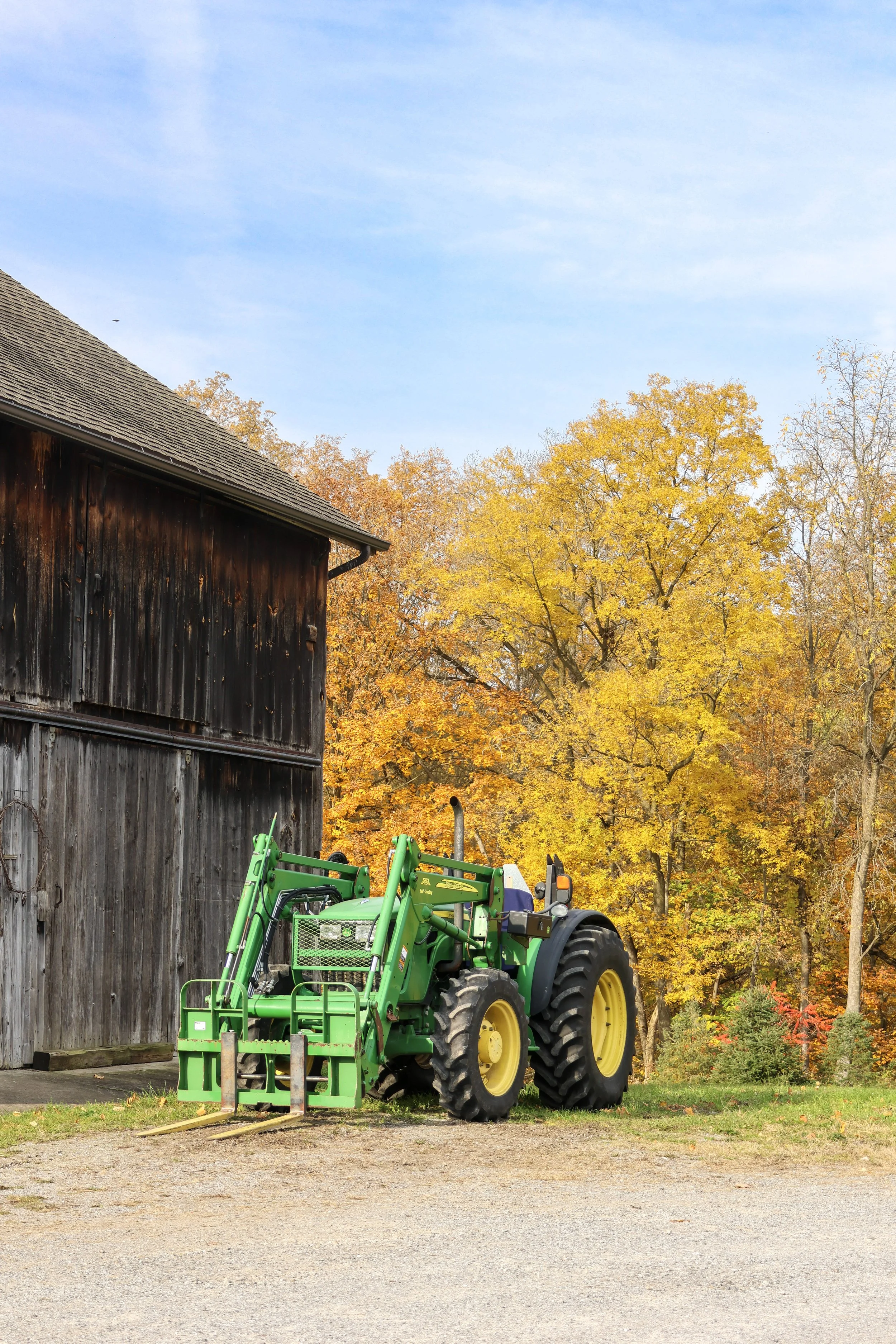 A farm scene with a green tractor parked on a gravel area near an old wooden barn. Trees with autumn-colored leaves are in the background under a blue sky.