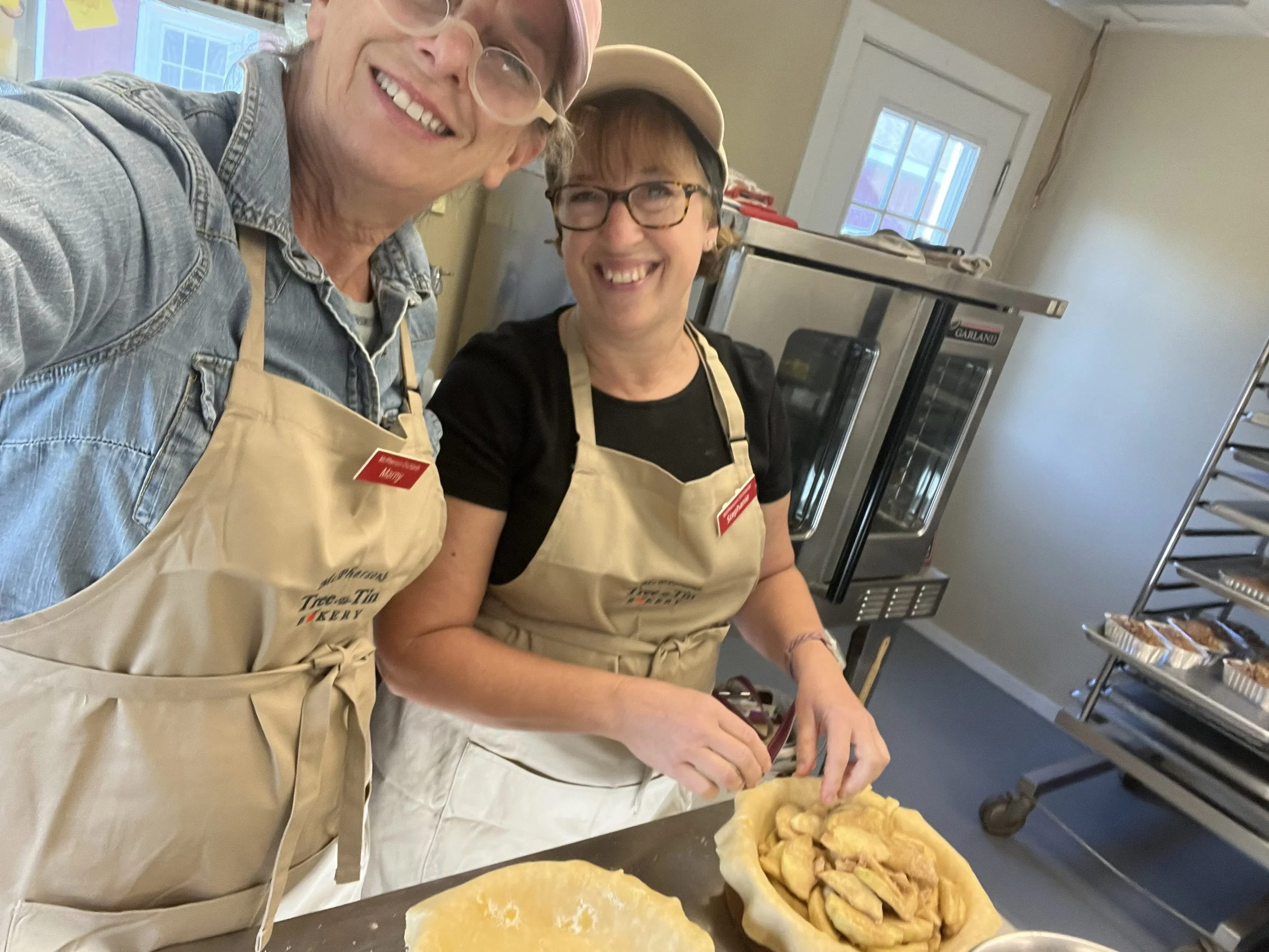 Two women wearing aprons and glasses in a kitchen, preparing food together. One woman is smiling and cutting something, while the other is laughing. They are surrounded by baking supplies and trays of dough or pastry.