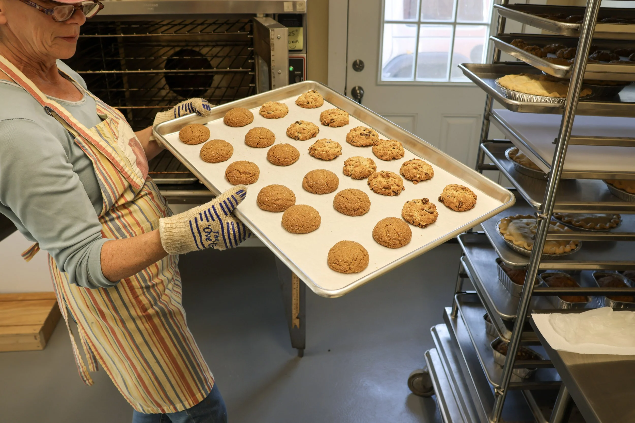 A woman wearing glasses, gloves, and an apron holding a baking sheet with cookies in a kitchen with racks of baked goods and a nearby window.