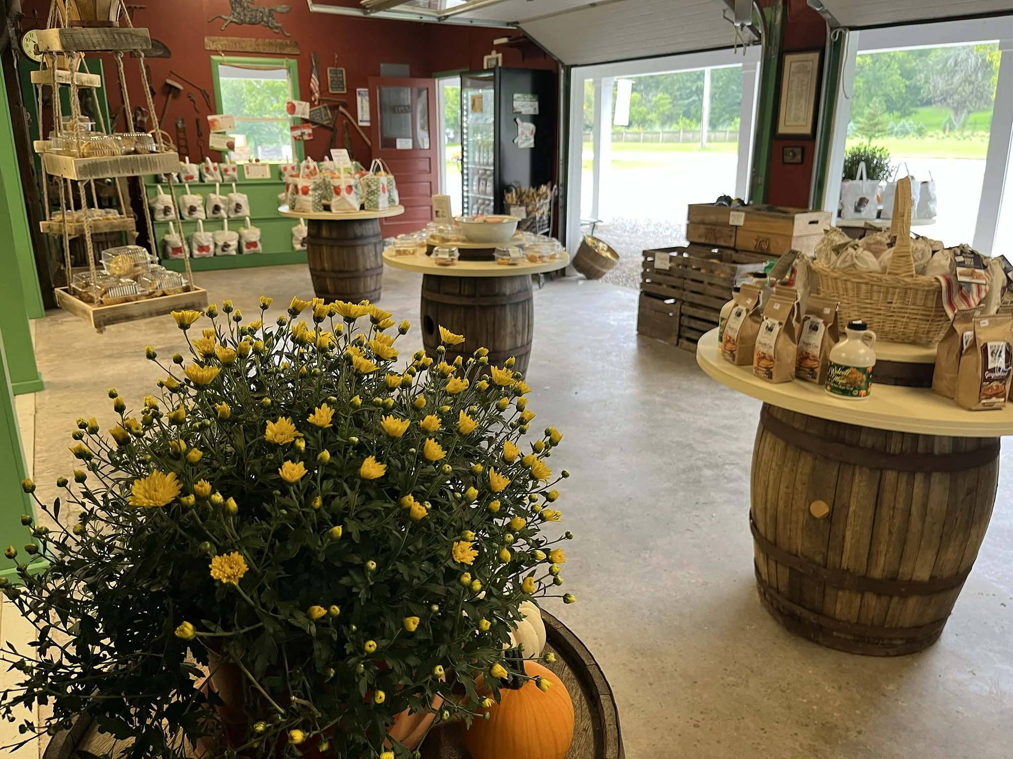 Interior of a shop with various items on display, including yellow flowers in a barrel, pumpkins, bags of food, and wooden crates, with open garage door revealing green outdoor scenery.