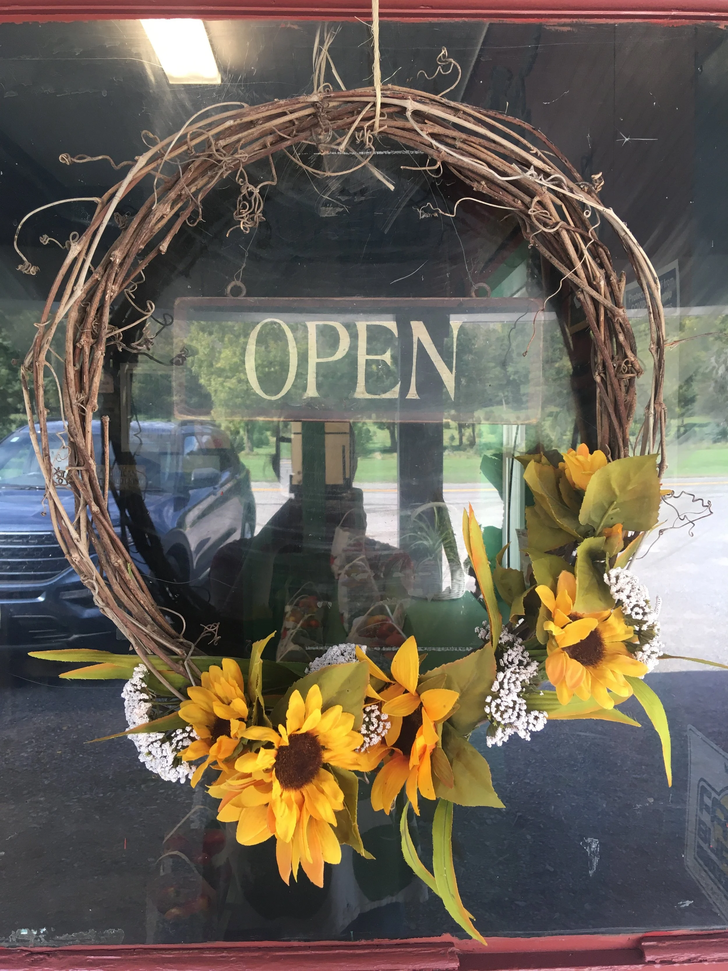 A floral arrangement featuring large yellow sunflowers with brown centers, complemented by small white filler flowers and green foliage, in front of a glass window with an open sign hanging behind it.