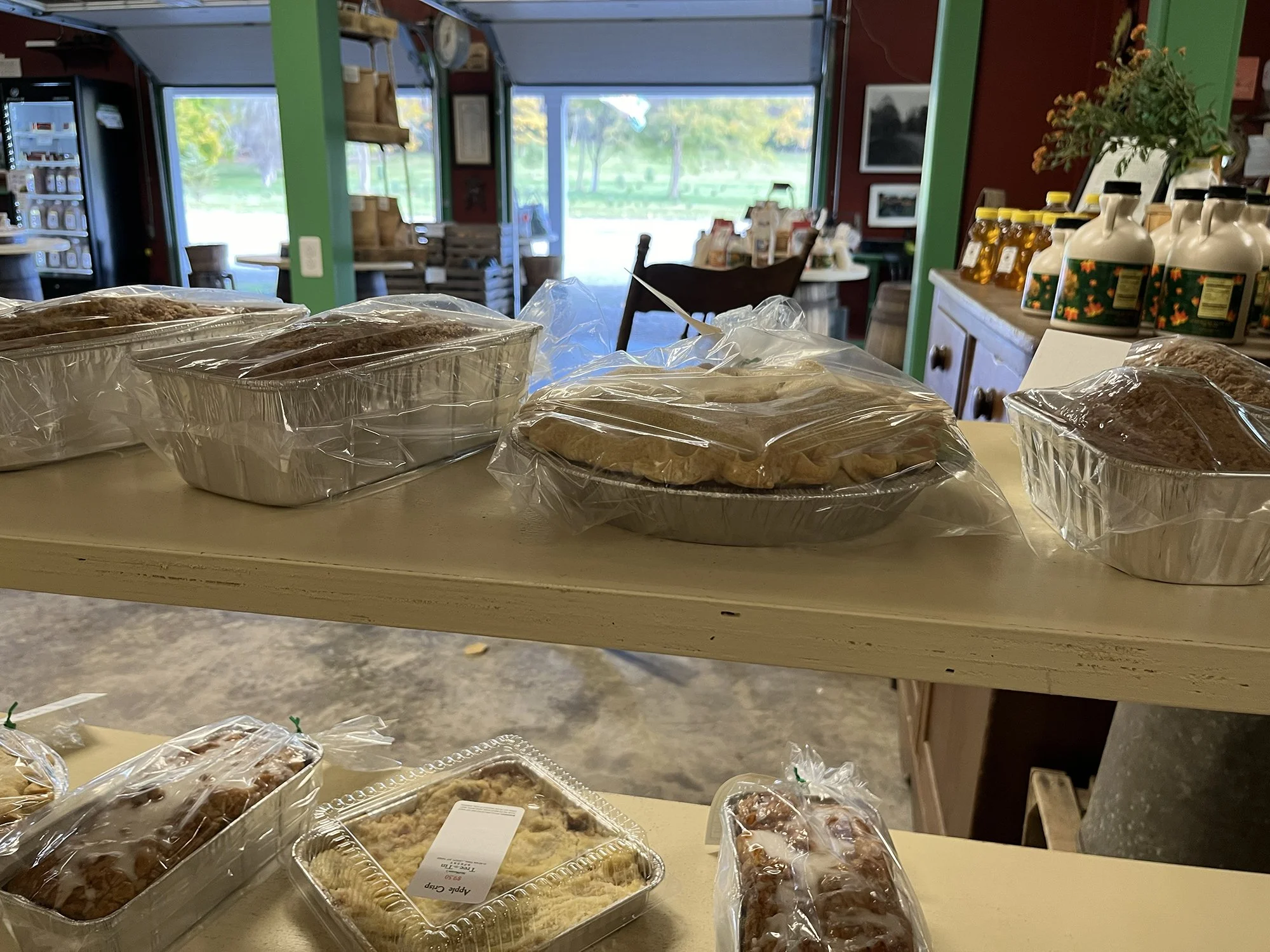 Display of baked goods in aluminum trays and plastic containers on a table in a cozy store with red and green walls, a wooden shelf, and a view of green trees outside.