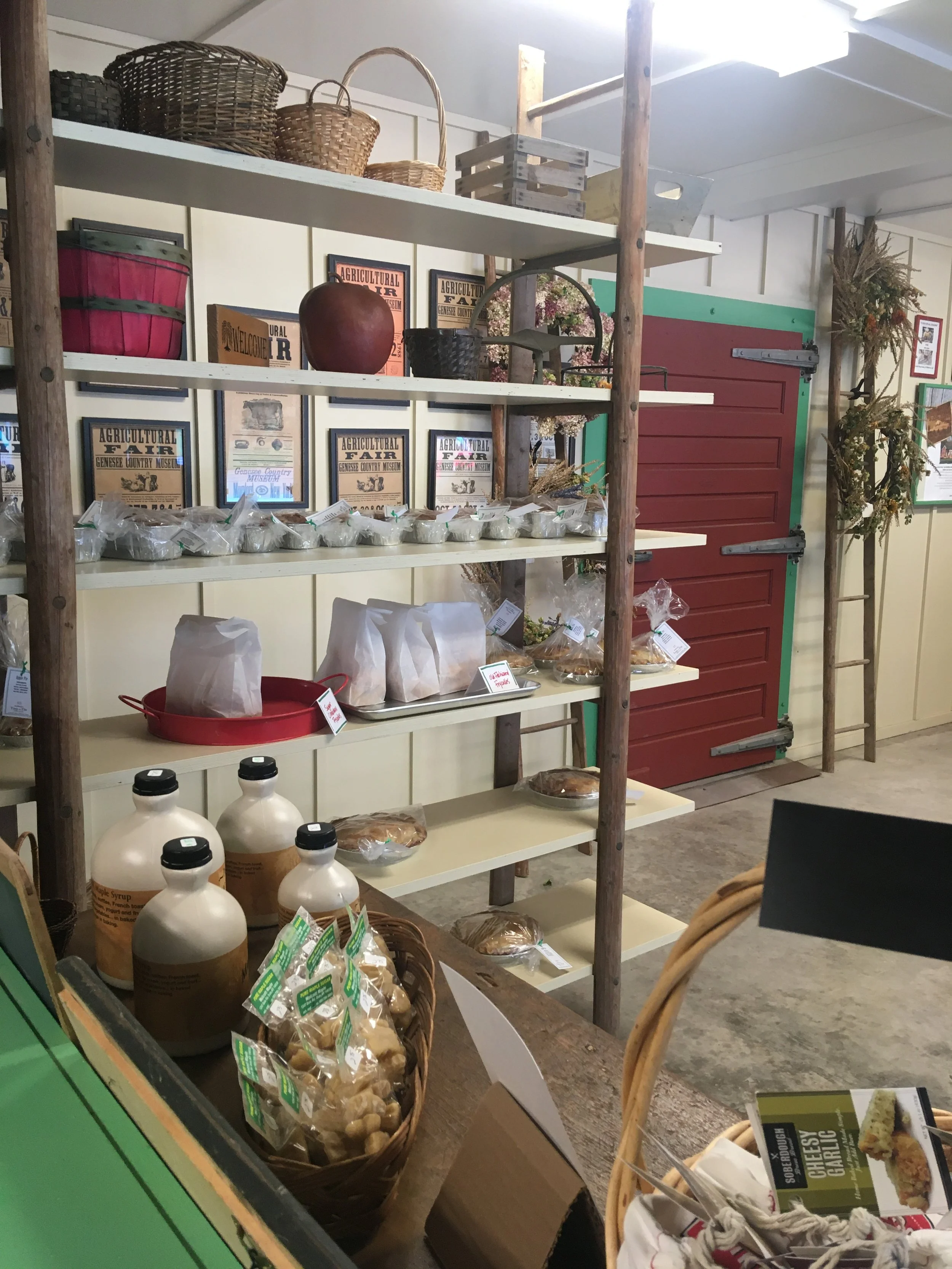 Display of farm-themed items including baskets, paper bags, and bottles on shelves and a table, with posters and door in the background.