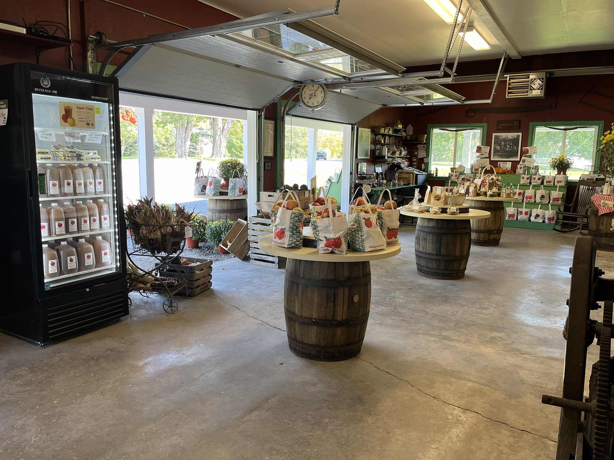 Interior view of a farmer's market with apple-themed products, including bags of apples on tables and a refrigerated display case with apple cider. Large windows reveal trees and a field outside, and the space is decorated with plants and framed pict