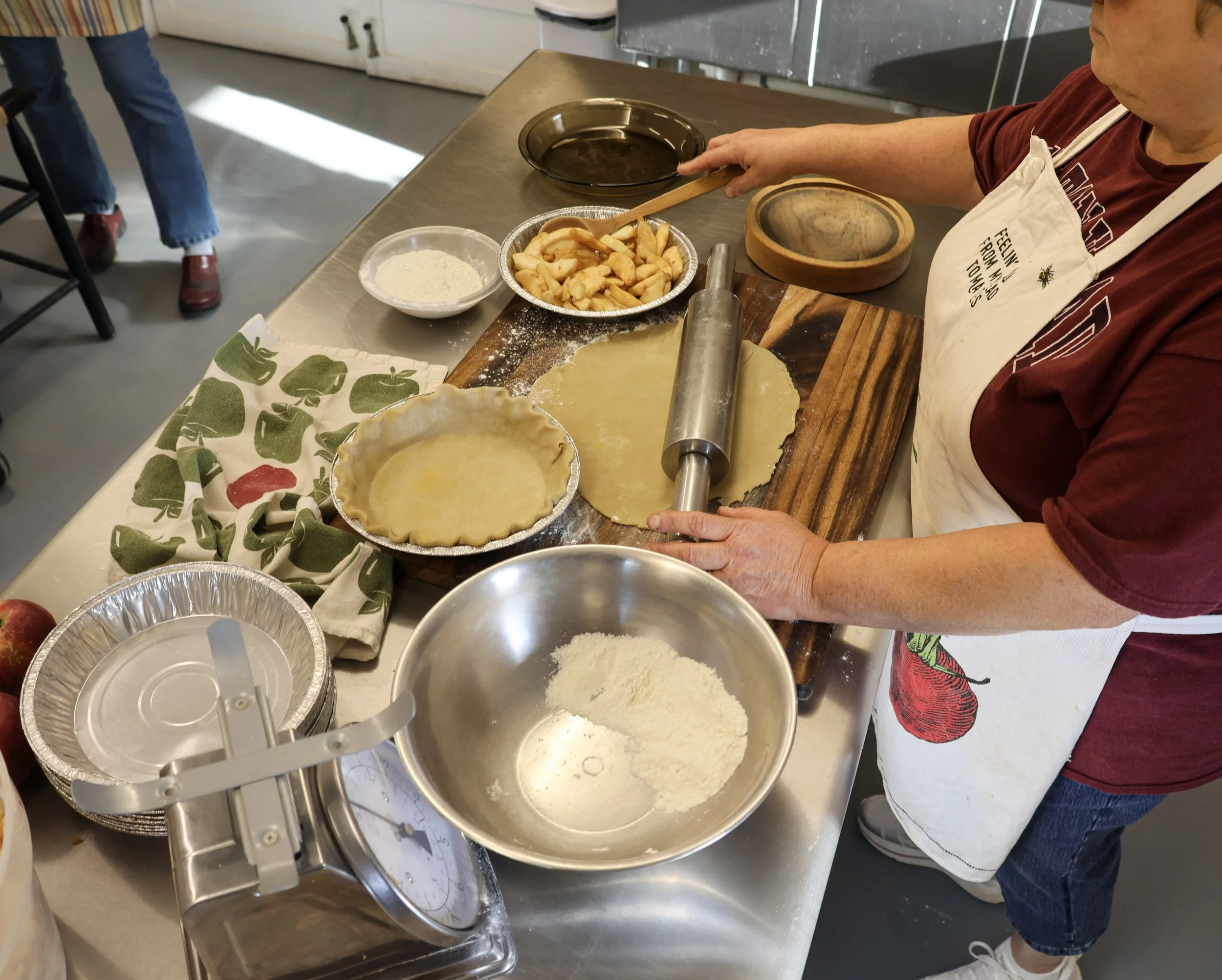 A person making pie crusts at a kitchen counter with bowls of sliced apples, flour, and a pie crust. The person is using a rolling pin and wearing an apron with a bee and the words 'Feeling from my toes to my nose' on it.