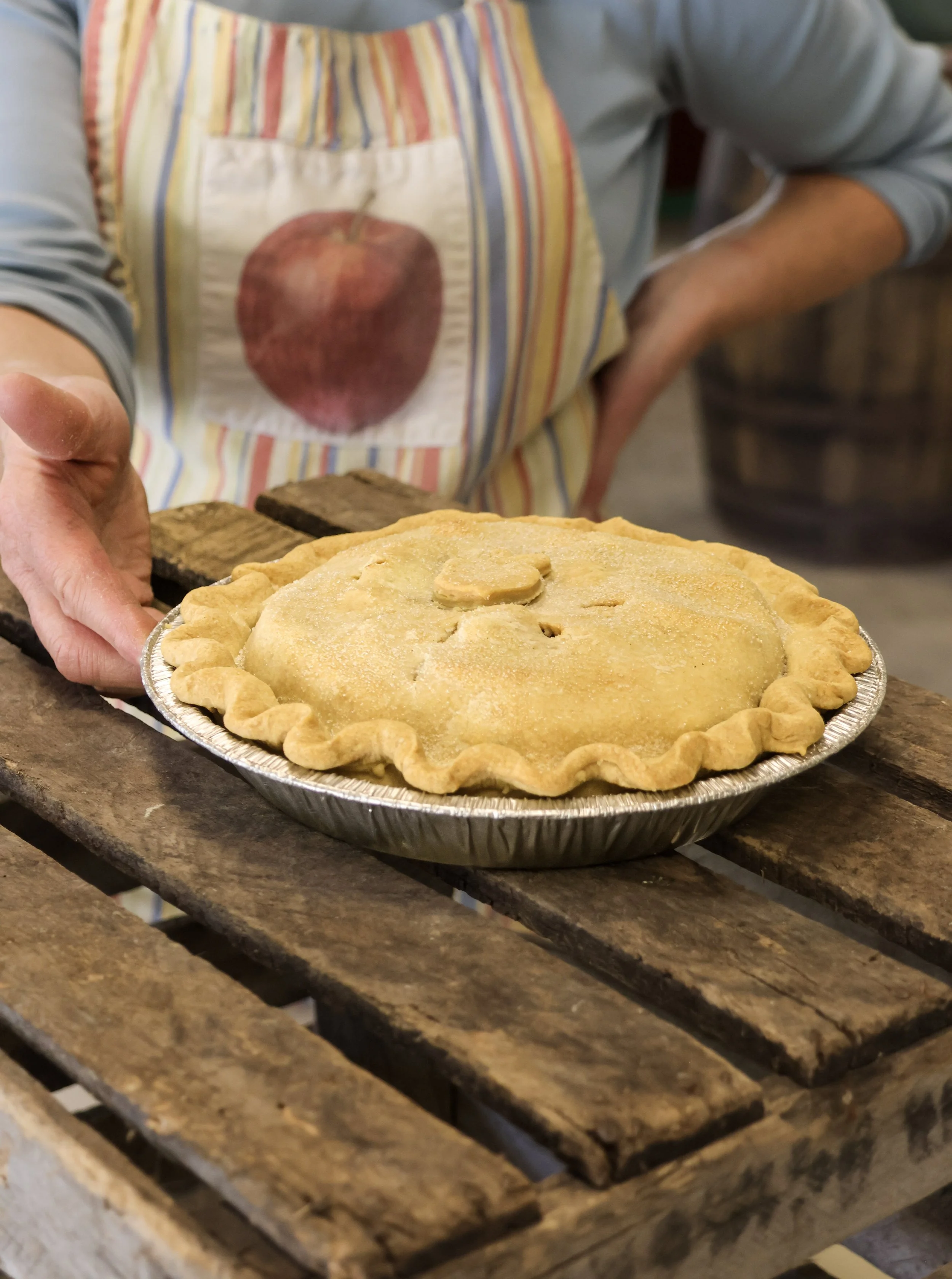 A person wearing a striped apron placing a homemade apple pie on a wooden table.