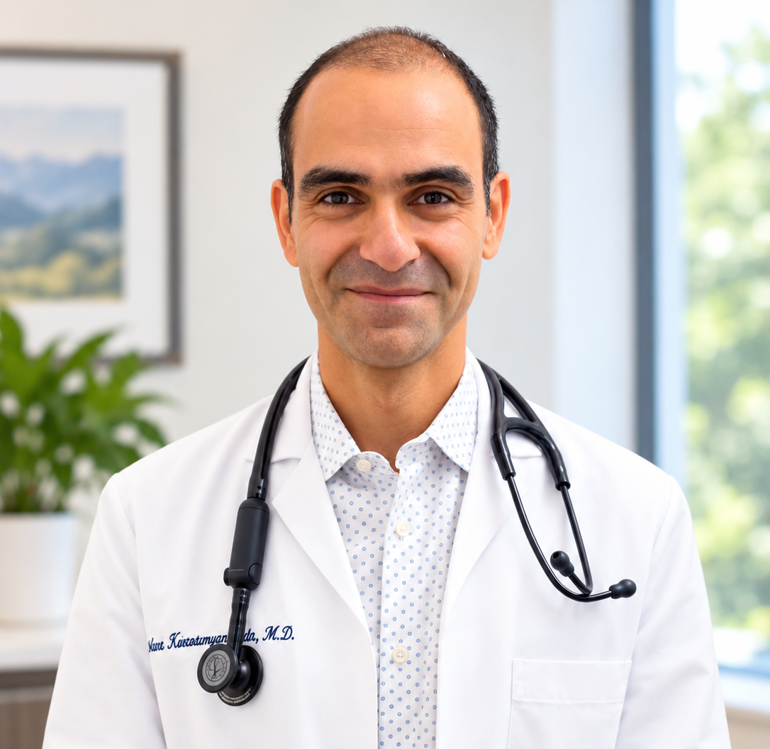 A male doctor with a stethoscope around his neck, wearing a white coat, smiling in an office setting with a plant and a window with greenery outside.
