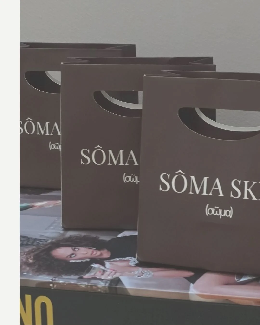 Three black shopping bags with cut-out handles and white text that reads 'SÔMA SKIN' with a small symbol underneath, placed on a table with a magazine or photo of a woman.