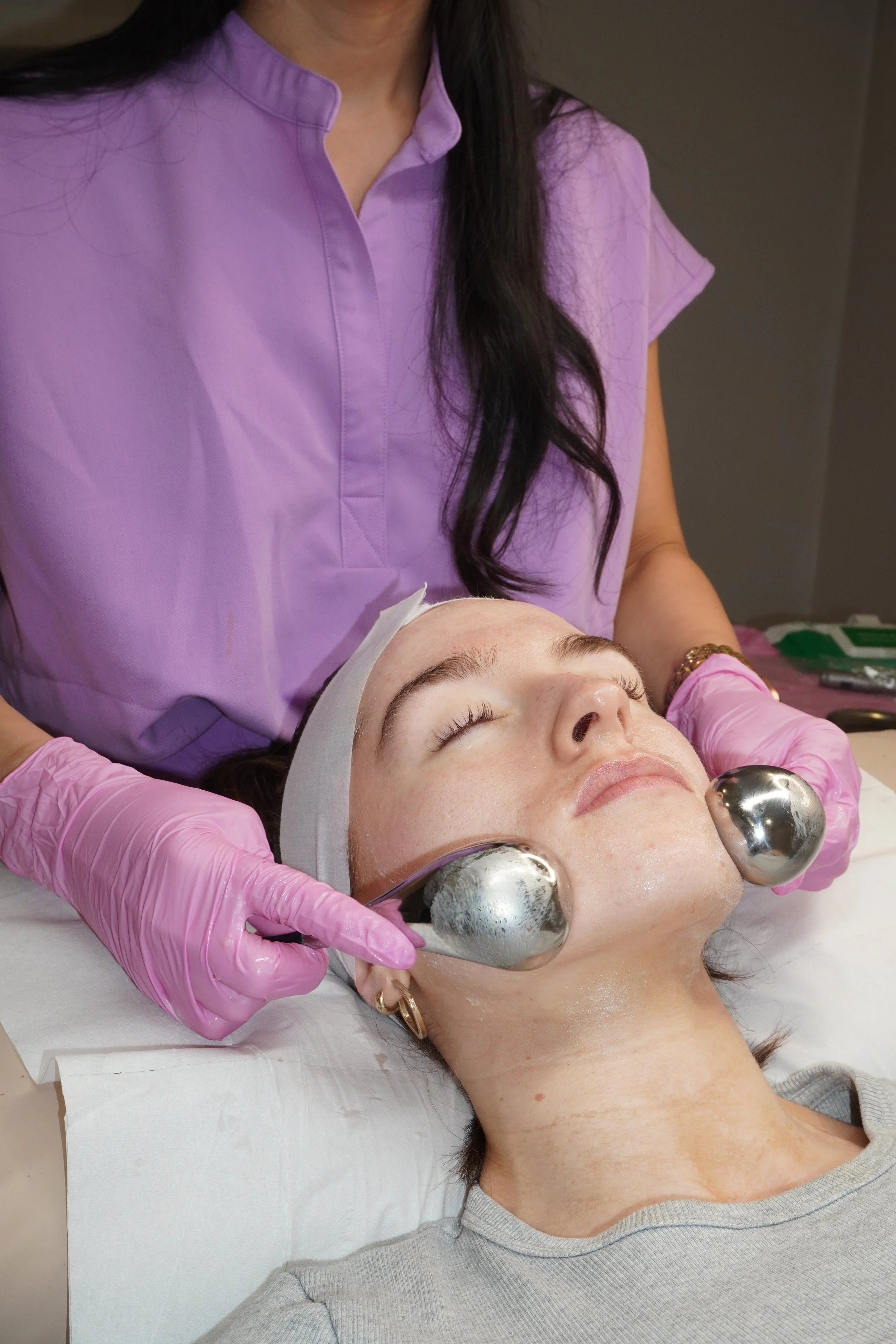 A woman lying on a treatment bed receiving a facial, with her eyes closed and wearing a headband. A skincare professional in a purple uniform and pink gloves is performing a facial treatment using tools with metal balls on her face.