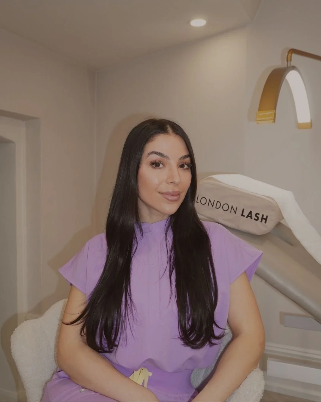 A woman with long dark hair sitting in a chair in a dental or beauty clinic, with a soft smile on her face, wearing a lavender outfit. Behind her is a dental or aesthetic treatment lamp labeled 'London Lash' on a beige cushion or neck rest.