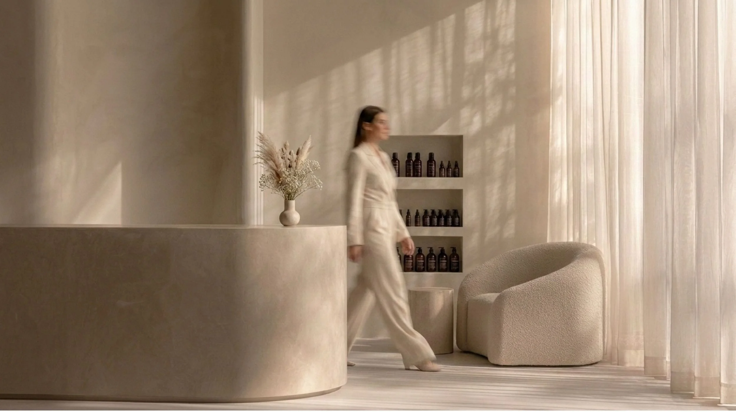 A woman in a beige outfit walking in a minimalist room with light-colored walls, sheer curtains, a built-in shelf with brown bottles, a vase with dried flowers, and a rounded beige armchair.
