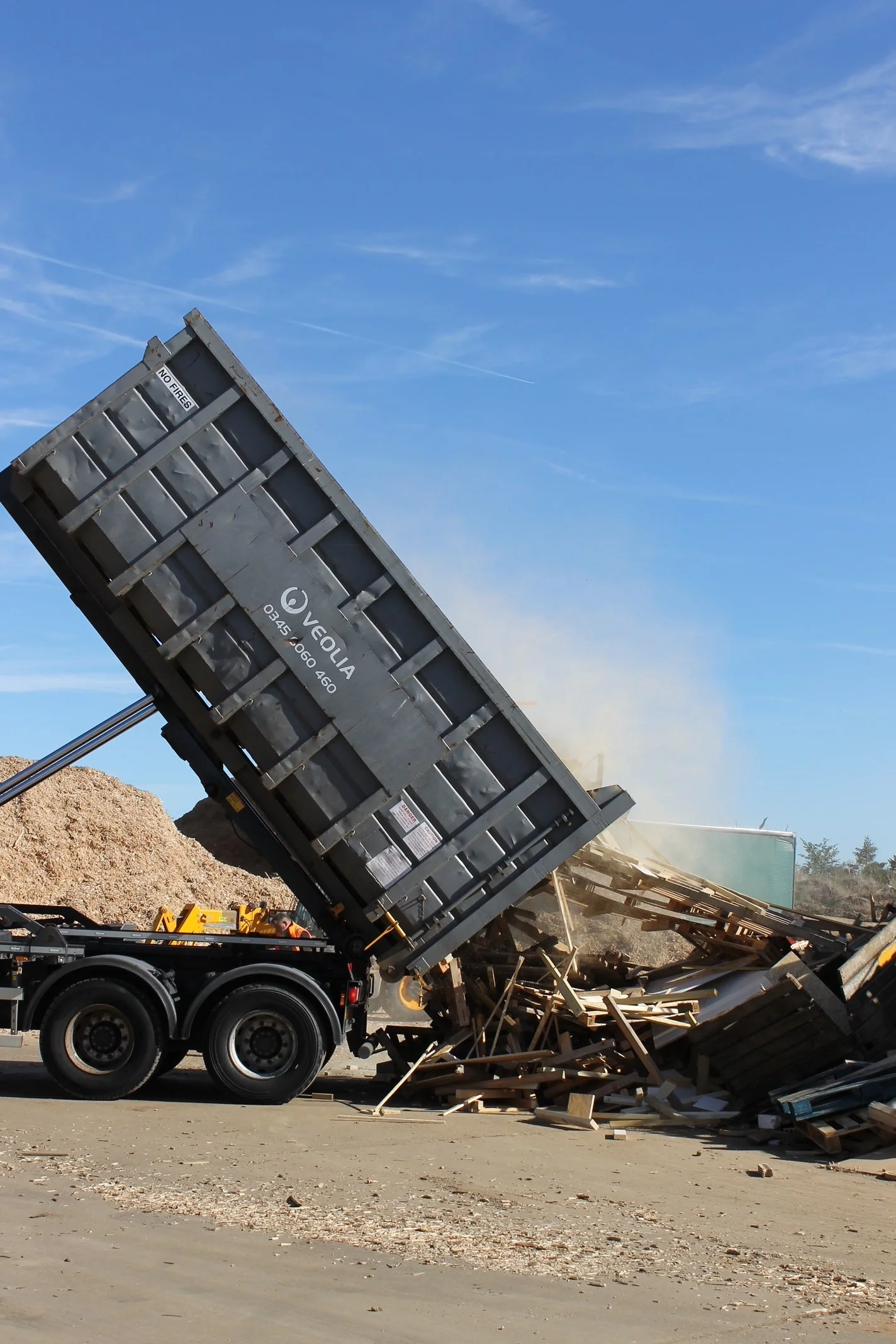 A dump truck lifting a bed filled with wood debris and broken wood pieces spilling out onto the ground at a construction or demolition site.