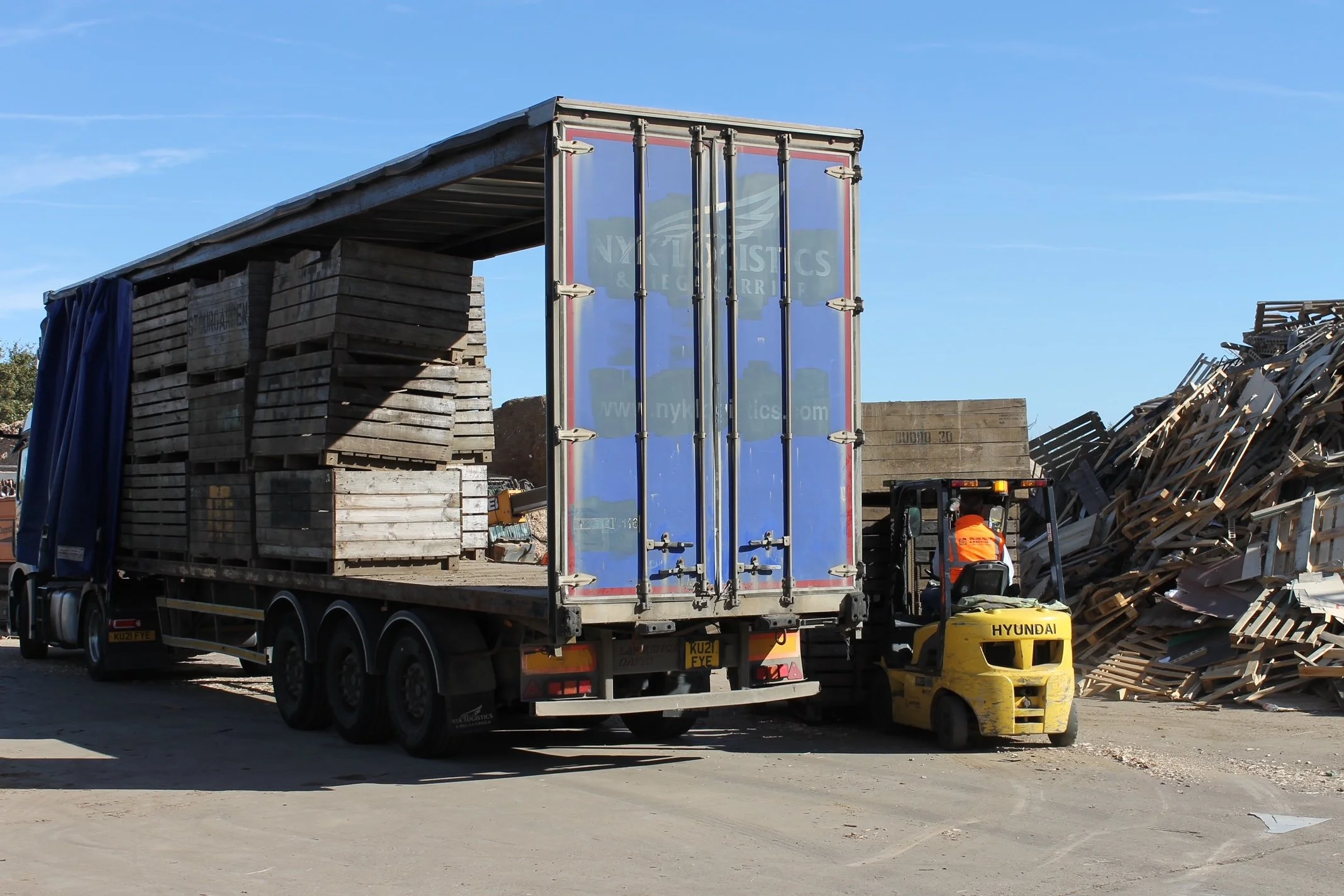 A large truck carrying stacked wooden pallets and a blue shipping container with the NYK Logistics logo, parked at an outdoor construction or junkyard site with a forklift and piles of wooden debris and scraps around.