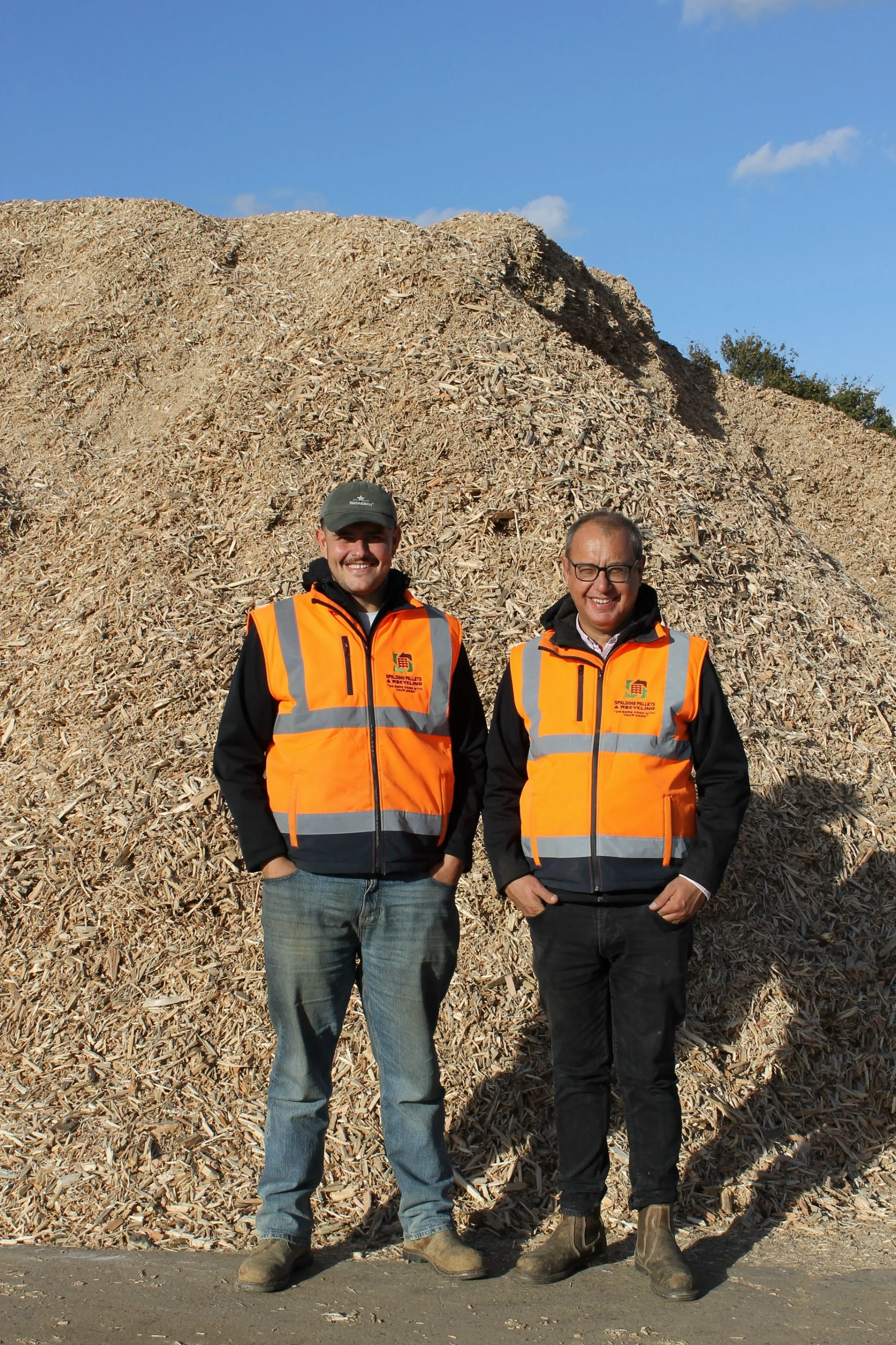Two men in orange safety vests standing outdoors in front of a large pile of wood chips, smiling, with a blue sky overhead.